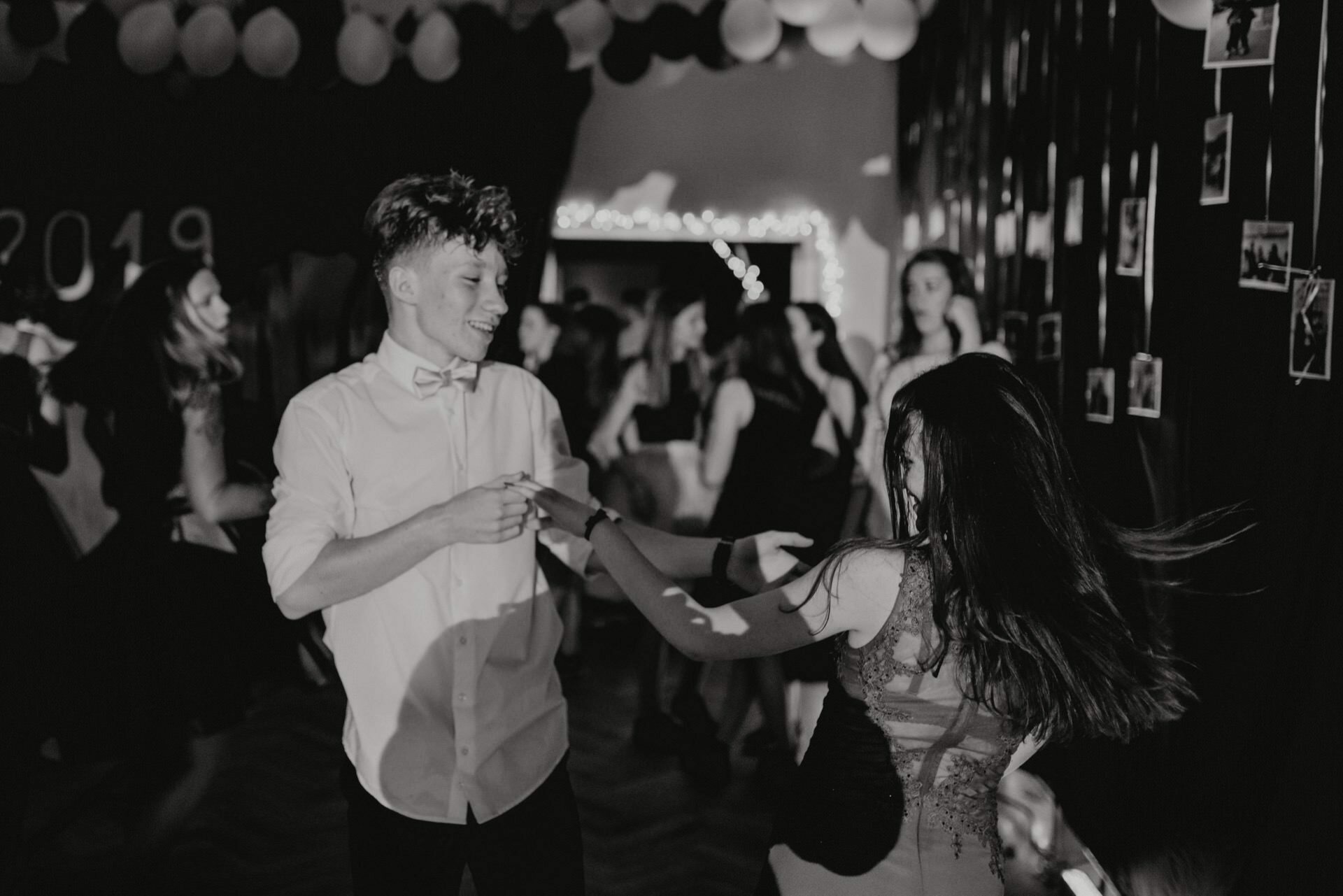 Black and white photo from a dance party. A young man in a white shirt and bow tie is holding hands and dancing with a young woman in a matching dress. They are surrounded by other dancing people, under the lights and balloons with the word "2019" on the wall in the background - a perfect photo of the event.  