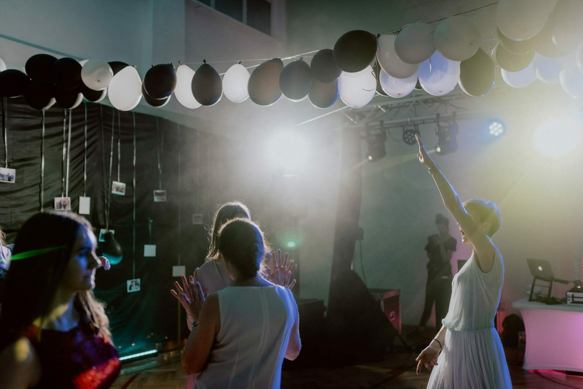 At the event, a group of people dance under a string of black and white balloons. The room, captured in an event photo report by event photographer warszawa, is dimly lit with colored lights. A DJ plays music in the background, and one person with short hair raises his hand and enjoys the rhythm.  