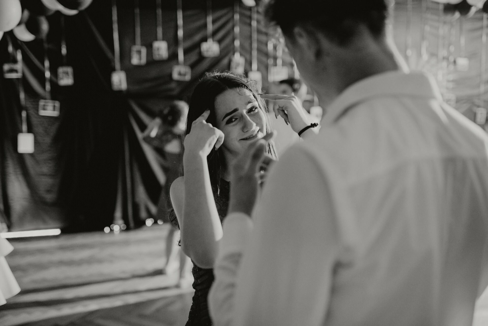 A black and white photo of a woman smiling and pointing her fingers at her temples while talking to a man. They're surrounded by decorations and other people in the background, creating a festive atmosphere - a perfect example of event photojournalism. 