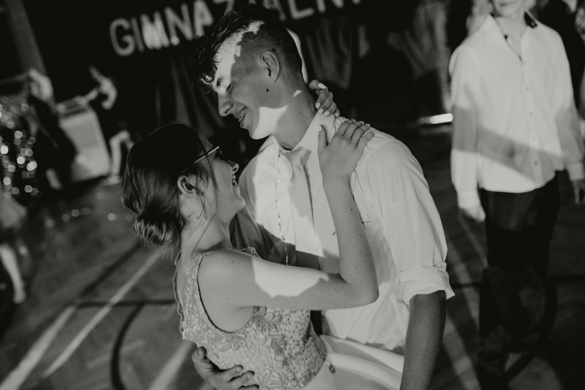 A young couple embracing and smiling as they dance together in a dimly lit room. The woman is wearing a sleeveless dress and the man a light-colored shirt. Other people are visible in the background, capturing a beautiful photo of the events with "GIMNA..." partially visible on the banner.  