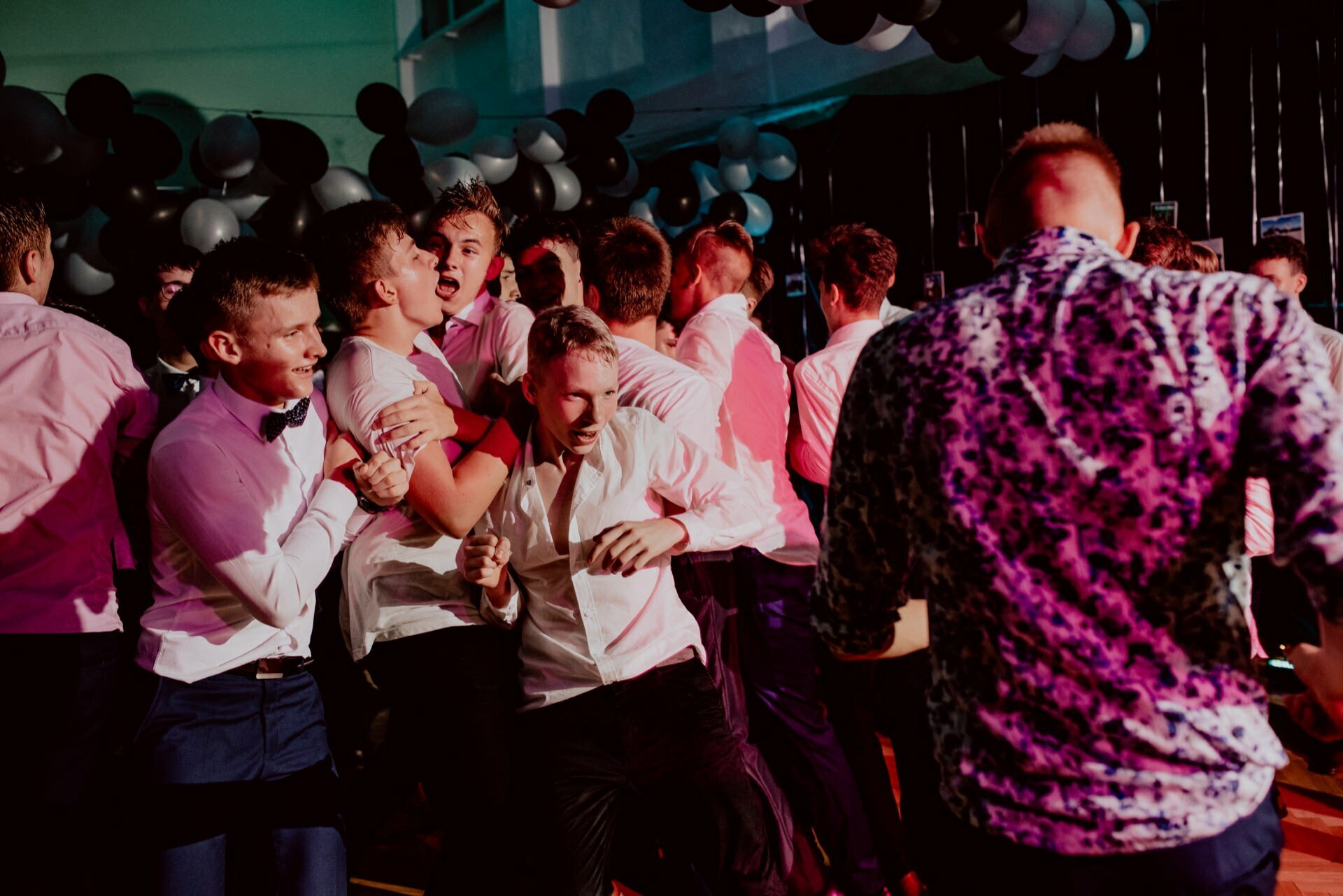 A group of young men dressed in shirts and pants laugh and dance enthusiastically during a lively party. The background is decorated with black and white balloons. One man wears a bow tie, while another wears a patterned shirt. The lighting is colorful - perfect for event photography to capture this vivid snapshot of the events.   