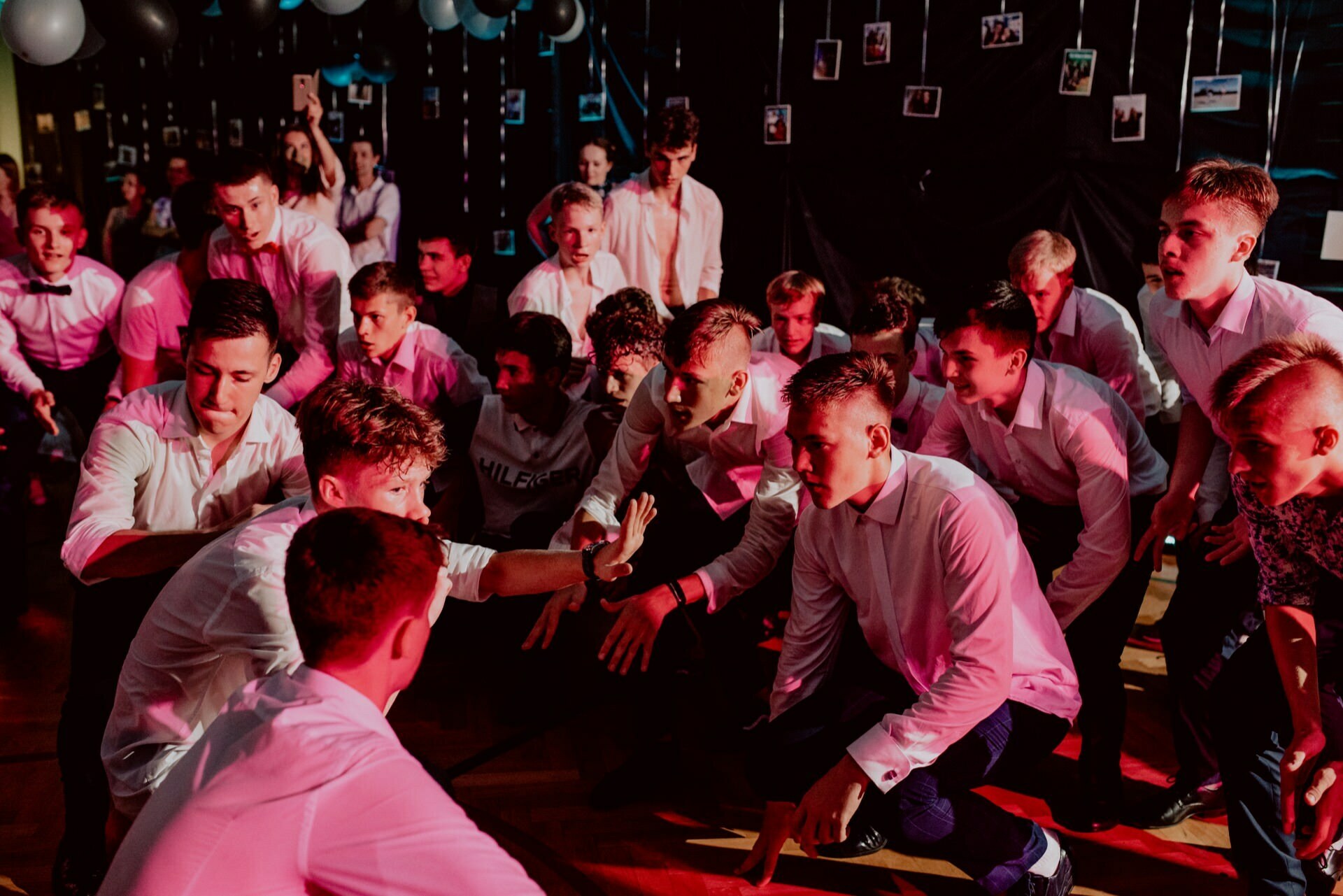 A group of young men in formal attire enthusiastically perform a dance routine during an event held indoors, against a backdrop of balloons and hanging photos. The lighting is red and pink, creating a festive atmosphere that was perfectly captured by the event photography. 