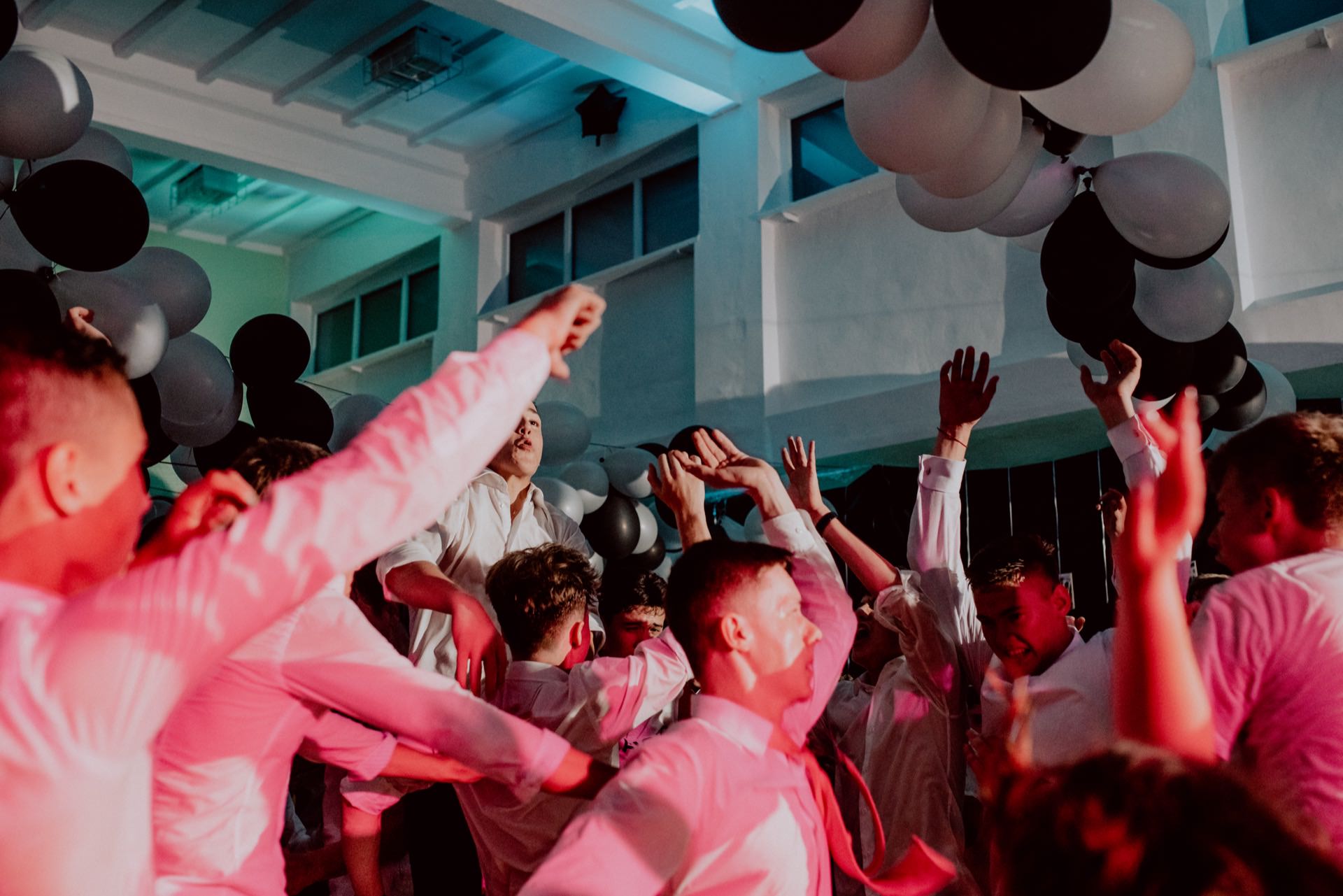 A group of young people in white shirts dancing and celebrating in a room decorated with black and white balloons. The lighting is vibrant and combines shades of red and blue to create a lively atmosphere. Some people have their hands raised in the air, which was beautifully captured by the event photographer warsaw.  