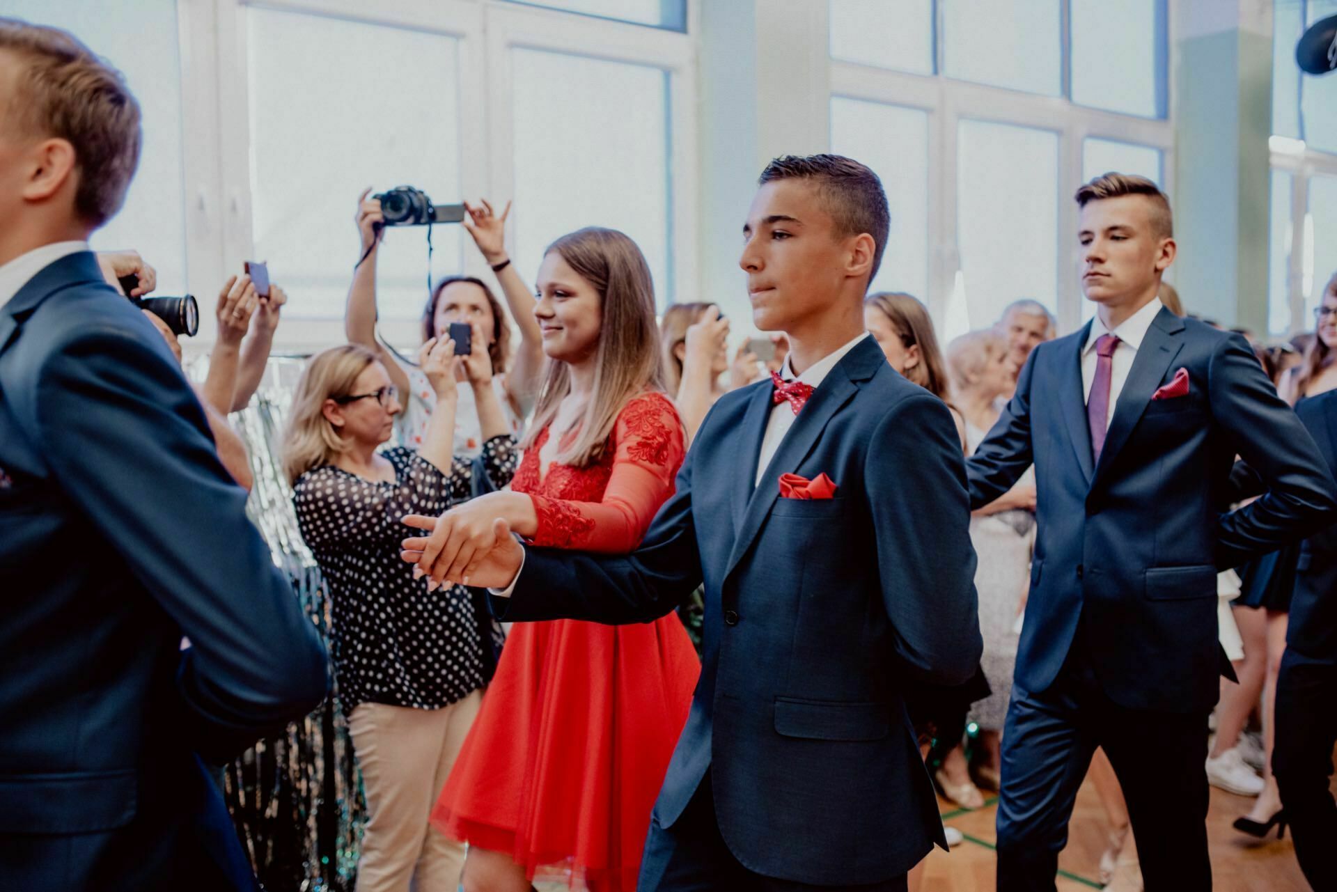 A young man in a suit and bow tie holds hands with a young woman in a red dress as they dance in a room full of people. Several spectators in the background take photos and videos of the event, capturing what could be an elegant photo essay of events at a formal social gathering or school event. 