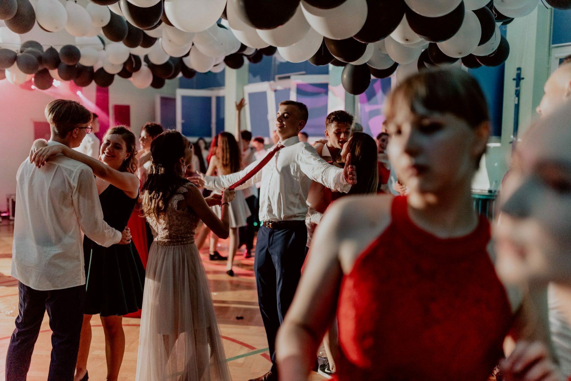 A group of young people dance at a decorated indoor party, beautifully captured by an event photographer Warsaw. They are dressed formally, some in dresses, others in suits. The ceiling is decorated with black and white balloons, and the lighting creates a festive atmosphere. The focus is on the dancing couples.   
