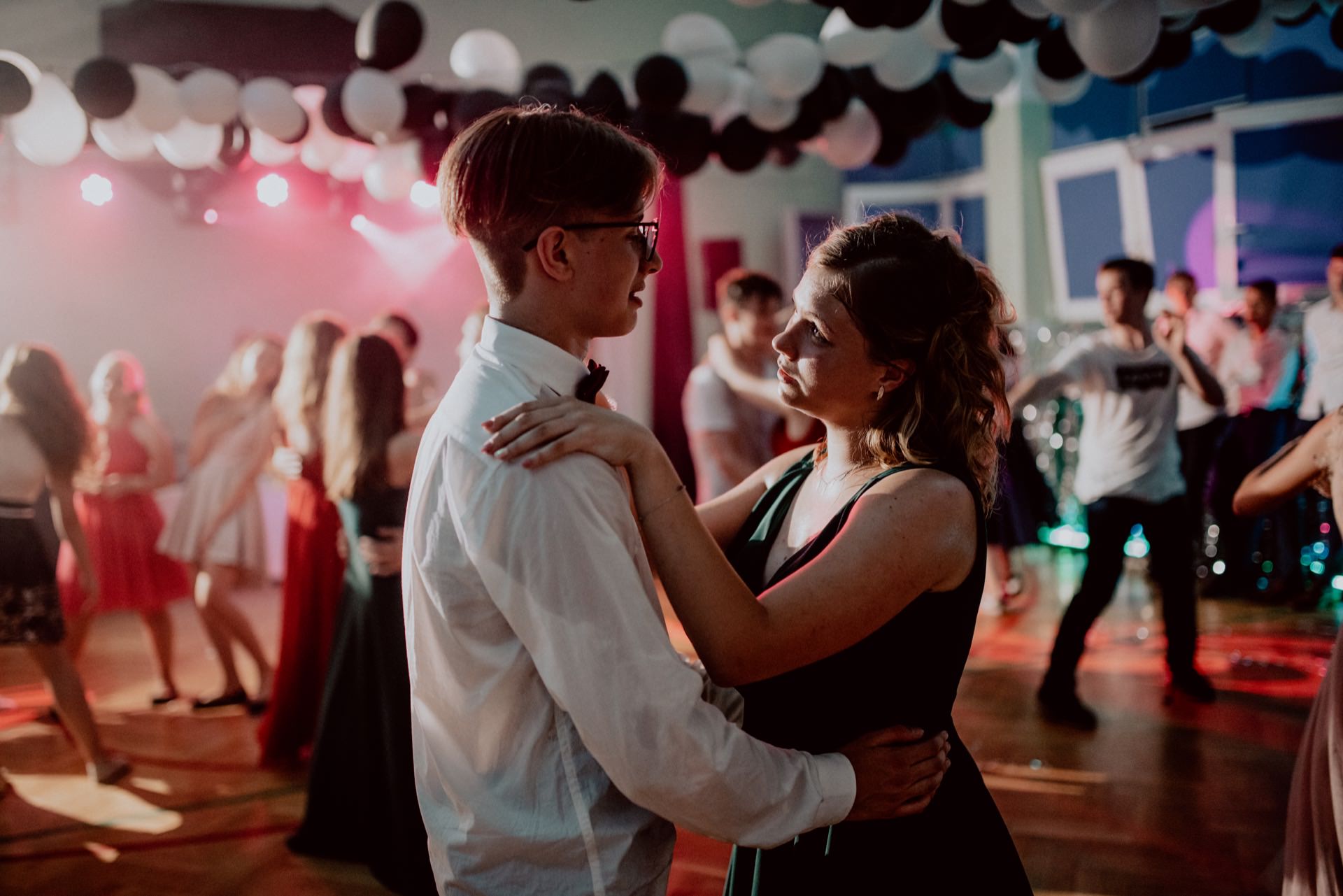 The bride and groom, dressed formally, dance close during the lively event with colorful lighting and balloons decorating the ceiling. Other people are also dancing in the background, creating a festive atmosphere - the perfect scene for any photography event. 