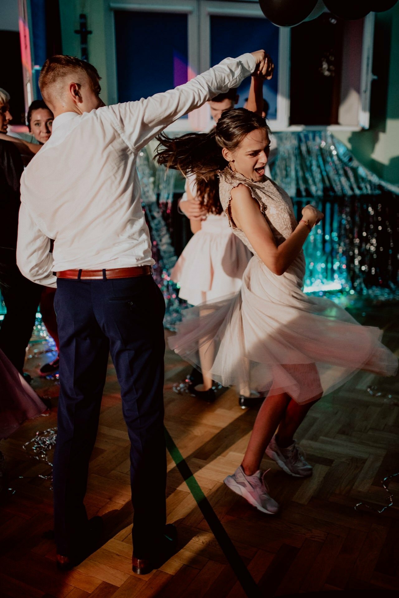 A bride and groom dance energetically at a party, the girl twirls around in an airy dress and tennis shoes, while the boy holds her hand. The room captured by the event photographer Warsaw is decorated with glittering candles and balloons, and several other people can be seen in the background. 