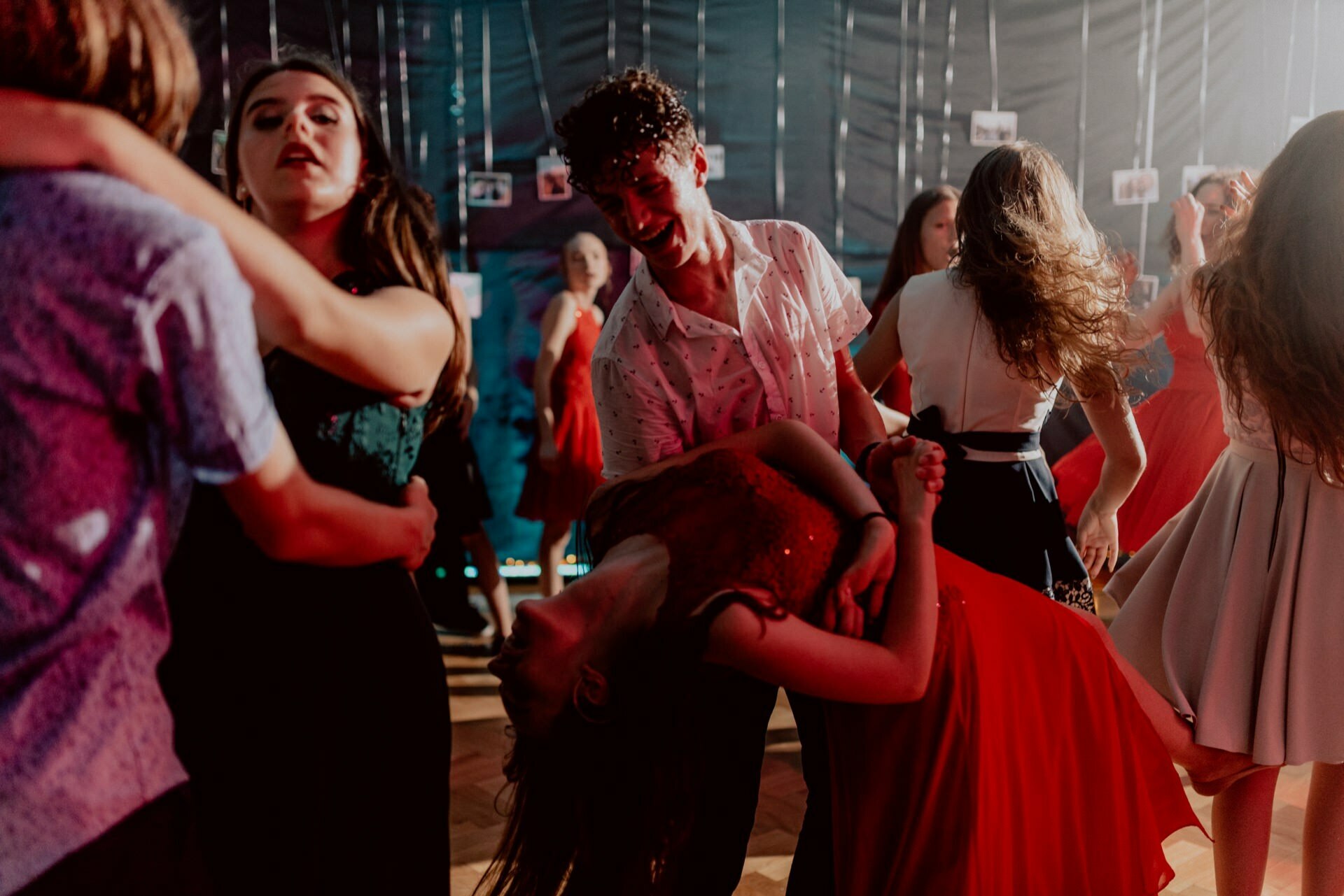 A group of young people are vigorously dancing in a dimly lit room. A couple dressed in white and red can be seen in the foreground, while a girl is being bent backwards by her partner. Others in different costumes dance around them, enjoying the lively atmosphere, which is perfectly captured by the event photography.  