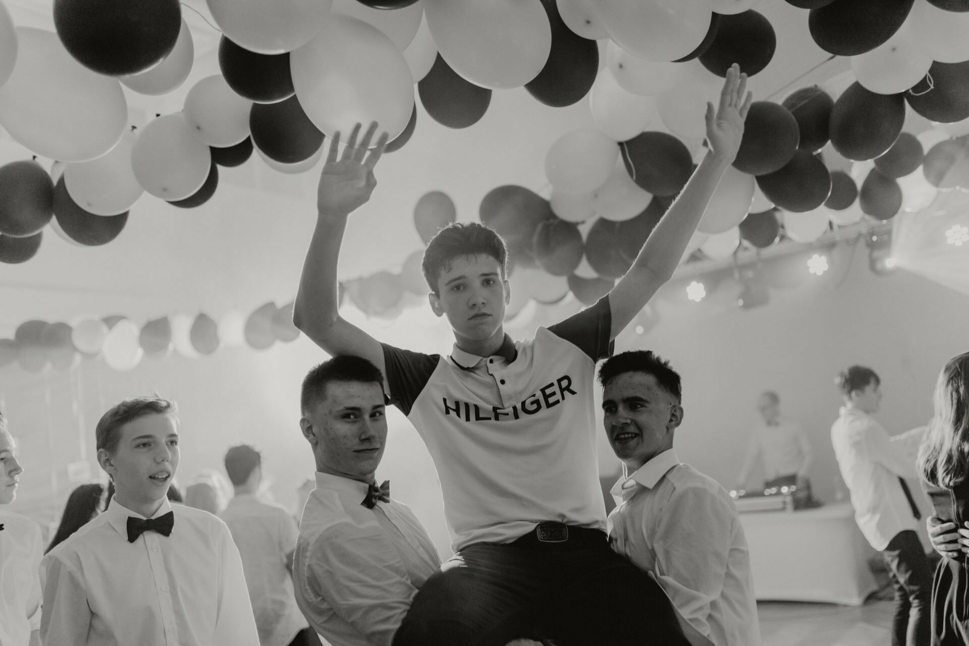 A group of young men celebrate at a party. One boy dressed in a "Hilfiger" shirt is carried over the shoulders of his friends with his arms raised triumphantly. They are surrounded by black and white balloons hanging from the ceiling, with more people and a DJ in the background - perfect event photography.  