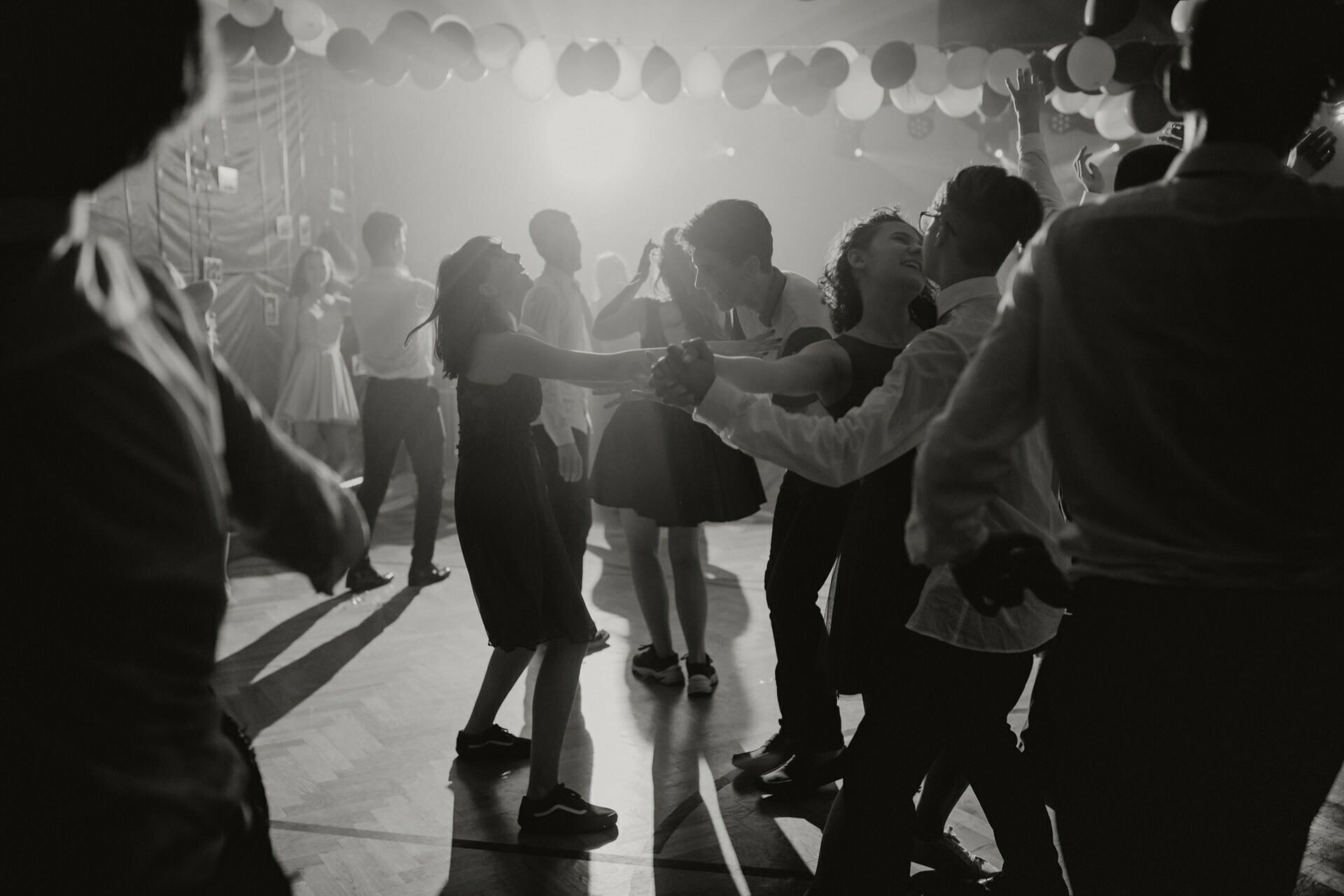 A black and white image of a bustling dance floor filled with people having fun. Several couples hold hands and dance energetically while balloons hang from the ceiling. The scene, captured with expert precision by event photographer Warsaw, is lit with soft, moody lighting, creating a festive atmosphere.  