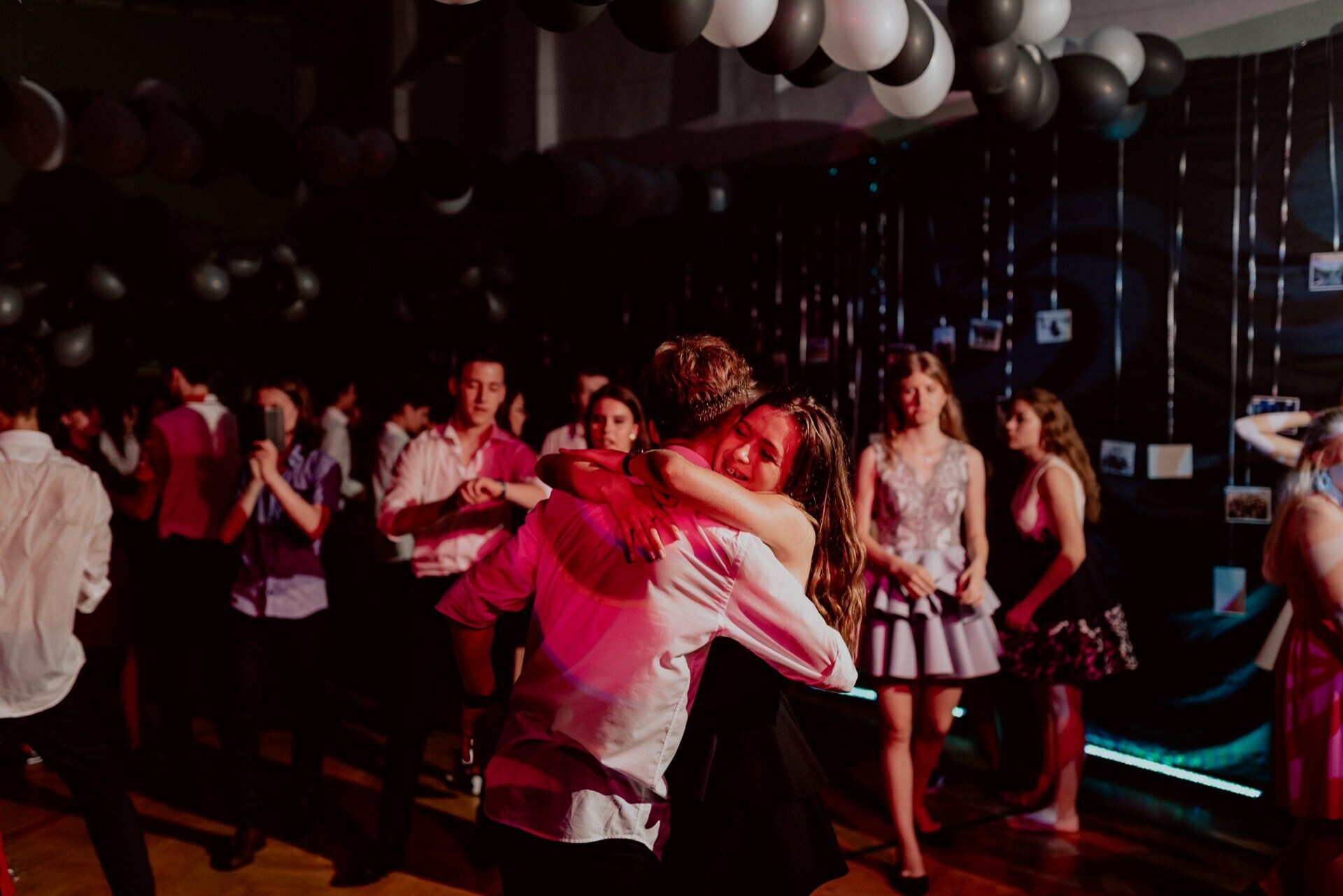 A dimly lit dance floor full of people dancing and mingling. In the foreground, a couple embrace close, while others move rhythmically in the background. The ceiling is decorated with black and white balloons, adding to the festive atmosphere captured by the event photographer Warsaw.  