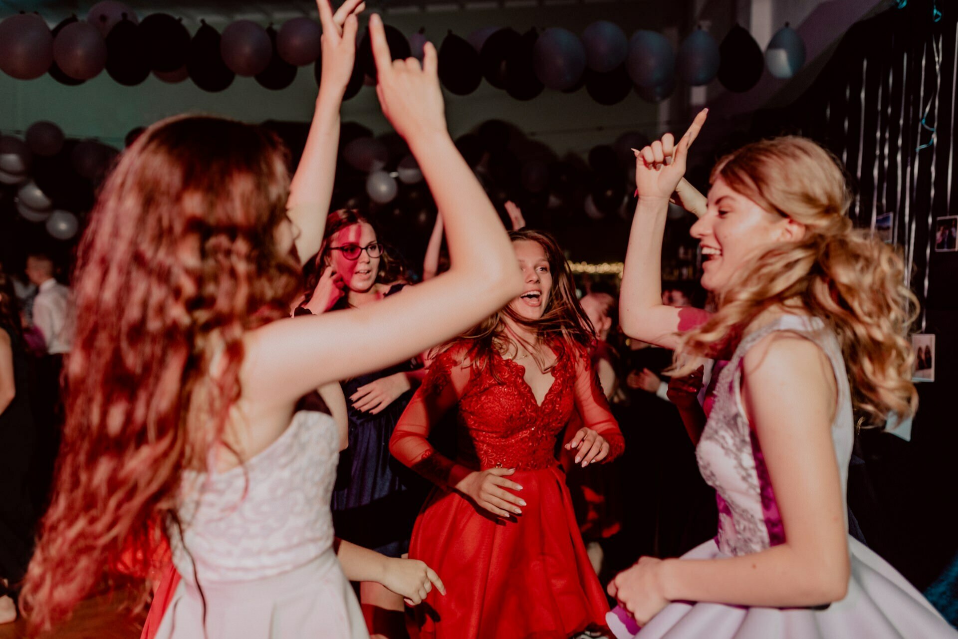 A group of girls dancing and having fun at a party, perfectly captured in a photo report of the events. They are dressed in formal attire, one wearing a red dress, others in different colors. With their hands up and smiles on their faces, they are surrounded by dim lighting and balloons.  