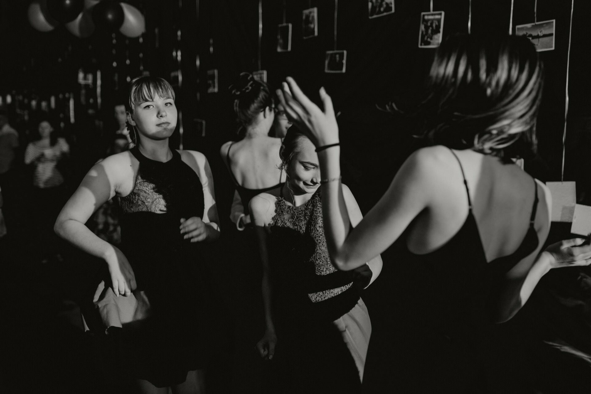 A group of young women in dark dresses dance and socialize at a dimly lit party, capturing the essence of event photography. In the background, photos hang on strings, and the left corner is decorated with balloons. The black and white image depicts a vivid, candid moment.  