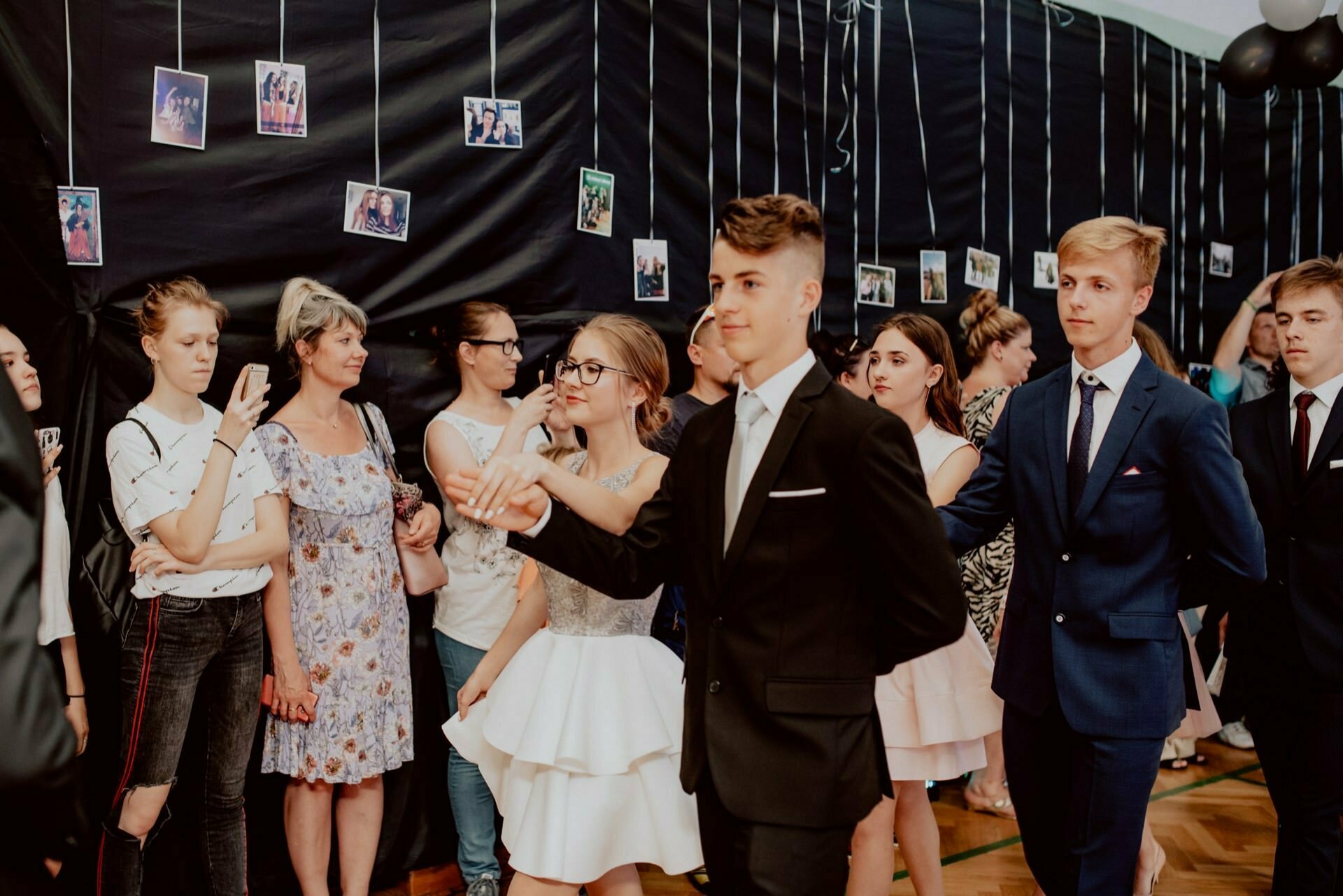 A group of young people in formal attire dance in a room decorated with hanging photographs. Carelessly dressed spectators stand against the walls and watch the dancers. The atmosphere seems festive and lively, perfect for event photography that captures every joyous moment in Warsaw.  