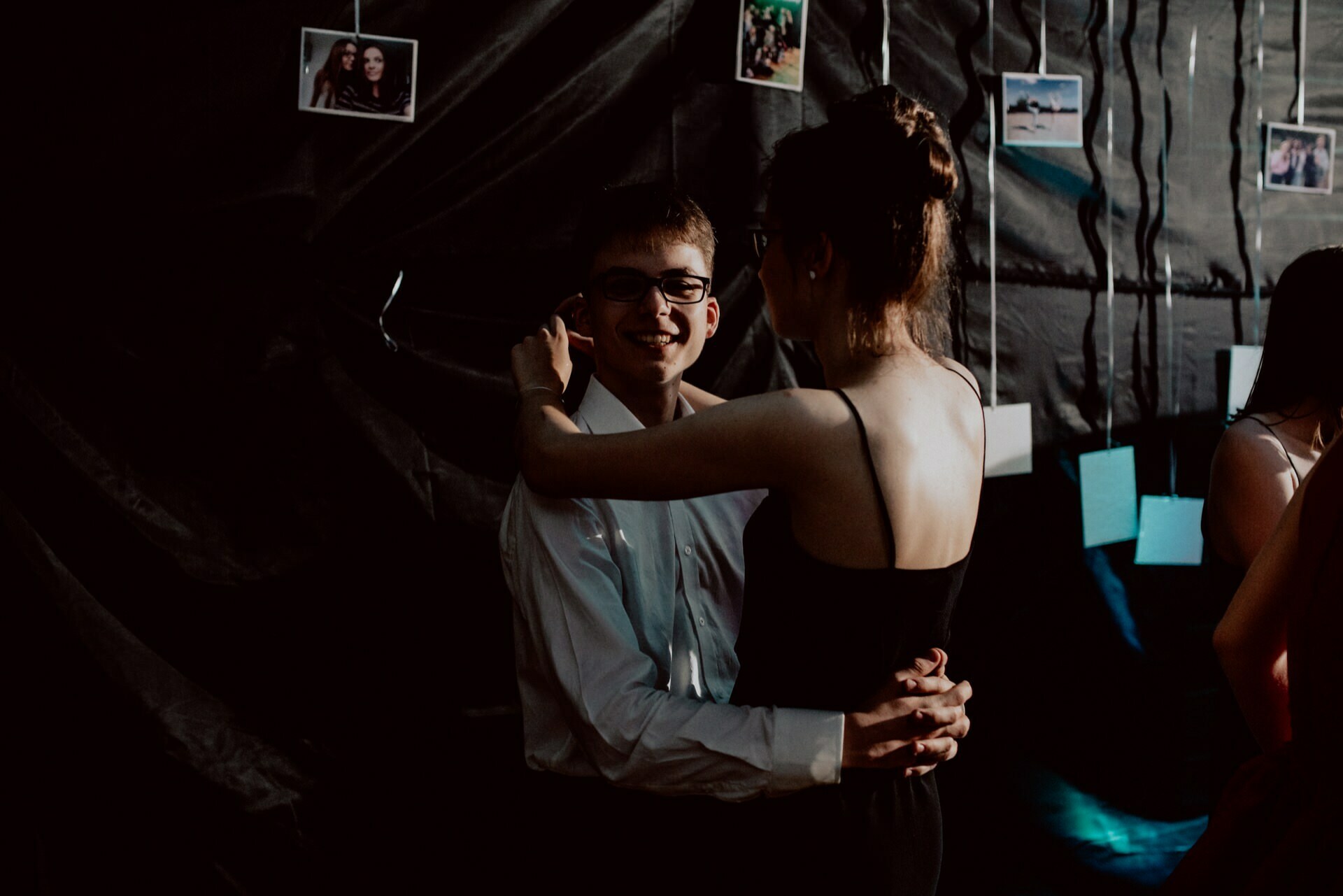 A young couple is dancing together at a dimly lit event. The man is wearing glasses, a white shirt and tie, while the woman is dressed in a black dress with her hair pinned up. Polaroid photos can be seen against a dark background behind them, capturing the essence of professional event photography.  