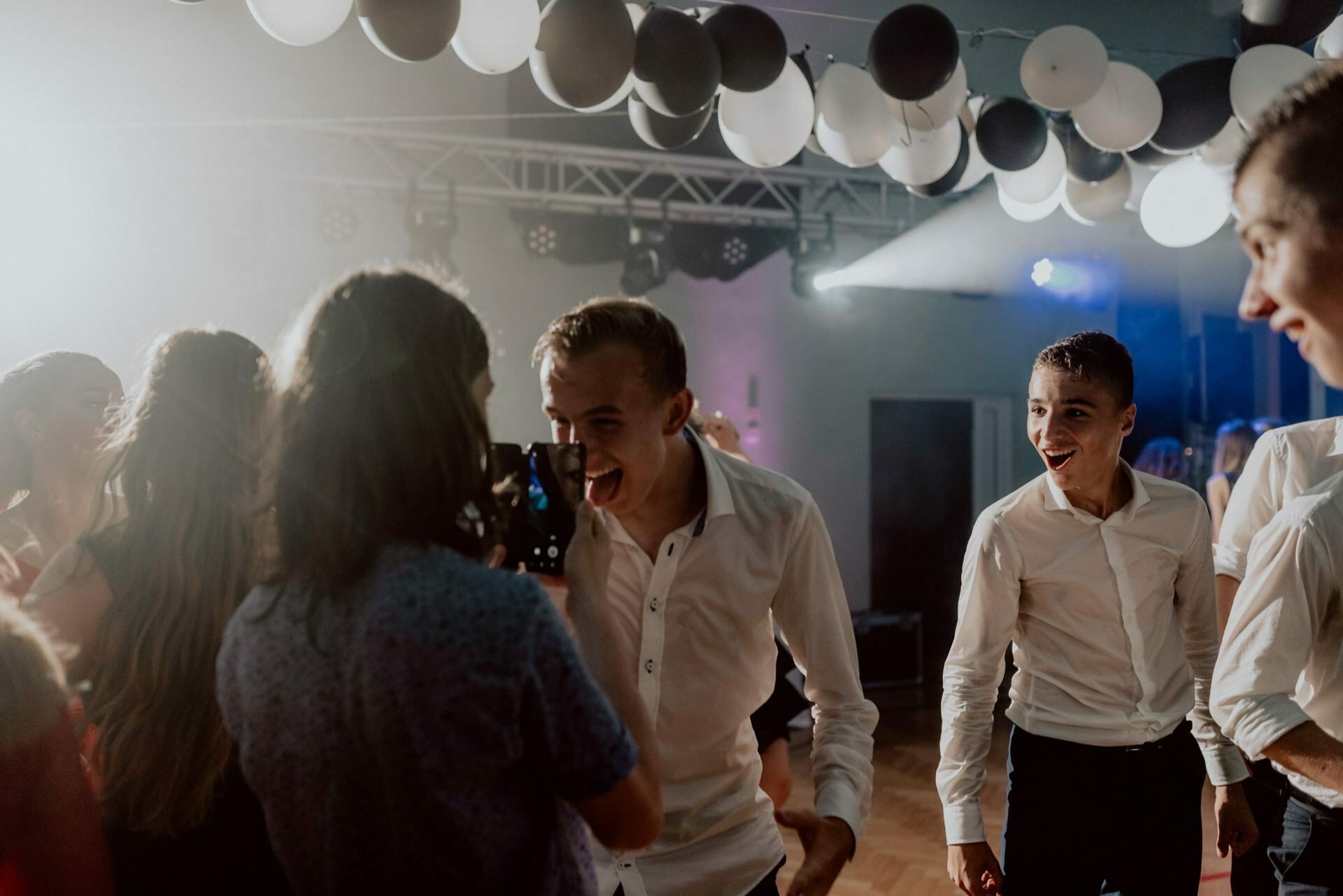 A lively group of young people dressed in semi-formal attire dance and party. The room, decorated with black and white balloons and illuminated by colorful lights, creates the perfect setting. A young man, smiling widely, holds his phone in the center, capturing memories for event photography.  