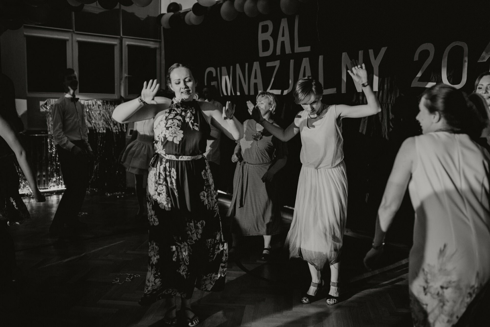 A group of people dancing merrily during what looks like a school event or prom. The setting consists of decorations and a background caption with the words "BAL" and "GIMNAZJAL 2024". The black-and-white photo captures the energy and movement, perfect for event photography by event photographer Warsaw.  