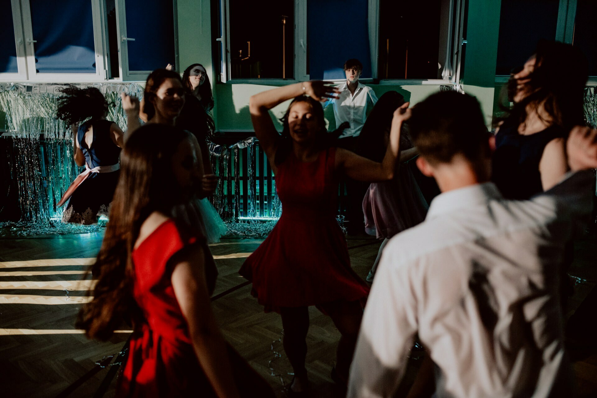 A group of people vigorously dance in a dimly lit room with colorful lights. The atmosphere seems lively and festive. Several people, mostly dressed in dresses, are in motion, enjoying the moment. The windows in the background suggest a room, ideal for event photography and photojournalism.   