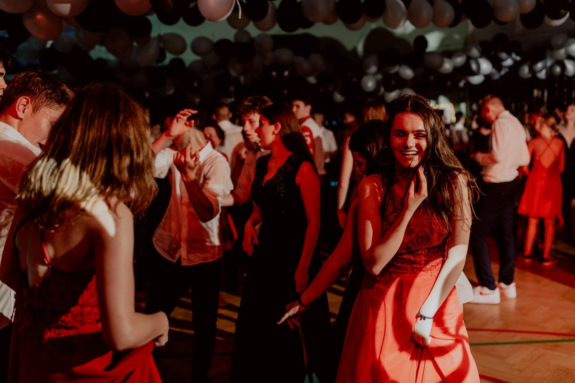 A joyful young woman in a red dress dances at a lively party under a ceiling filled with black and white balloons. She is surrounded by other guests who are also dancing and enjoying the event. The room is bathed in red and dim light, which adds to the festive atmosphere beautifully captured in this event photography.  