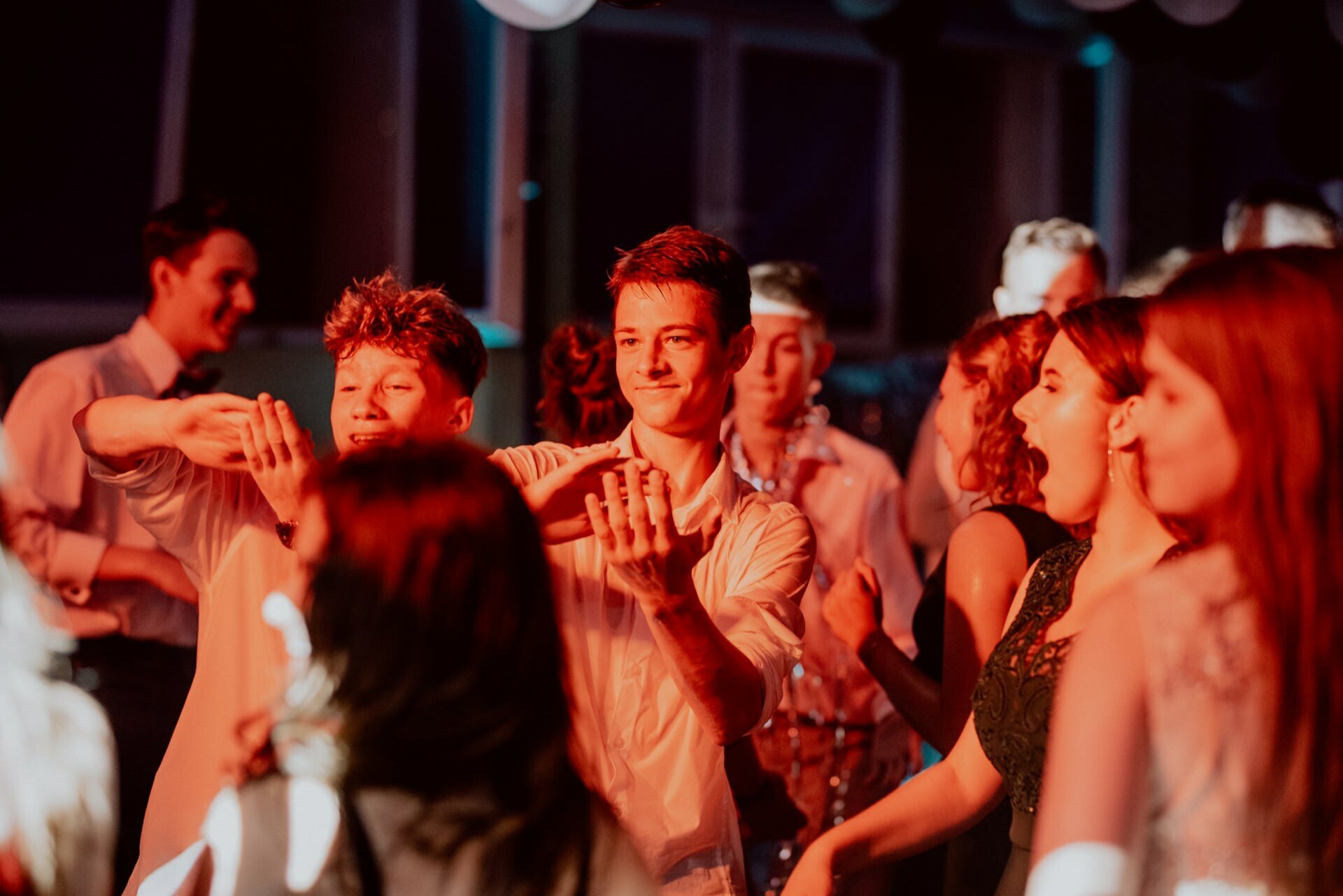 A group of young adults dancing vigorously in a dimly lit room with red lighting. Some are laughing, others are throwing their hands up in the air. The mood is joyful and lively, perfectly captured by the event photographer warsaw in the photo report of the events.  