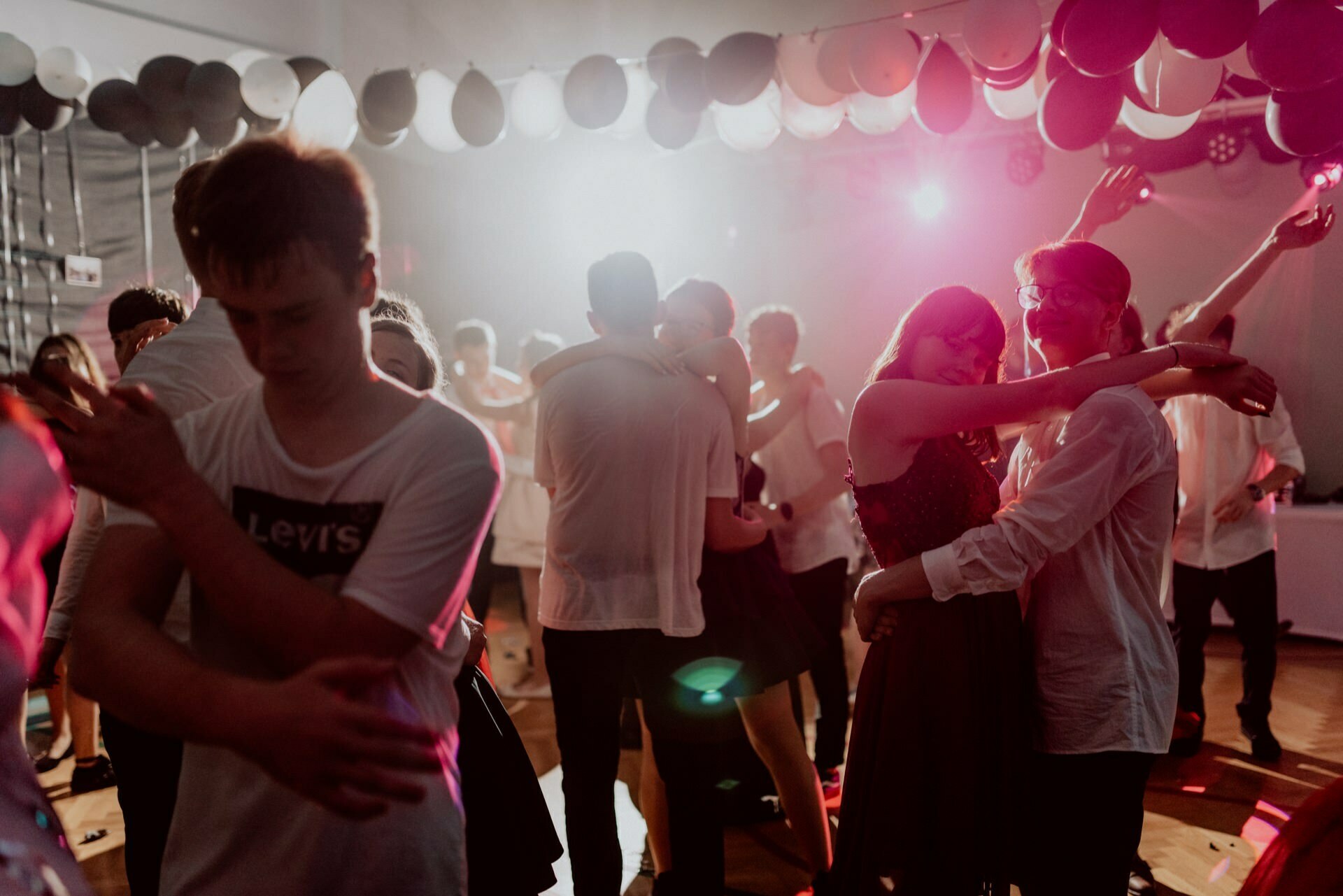 A group of people dance close at a party under a ceiling decorated with black and white balloons. Dim lighting combined with colorful spotlights creates a lively and festive atmosphere. One person in a white Levi's t-shirt stands out in the foreground, perfect for event photography.  