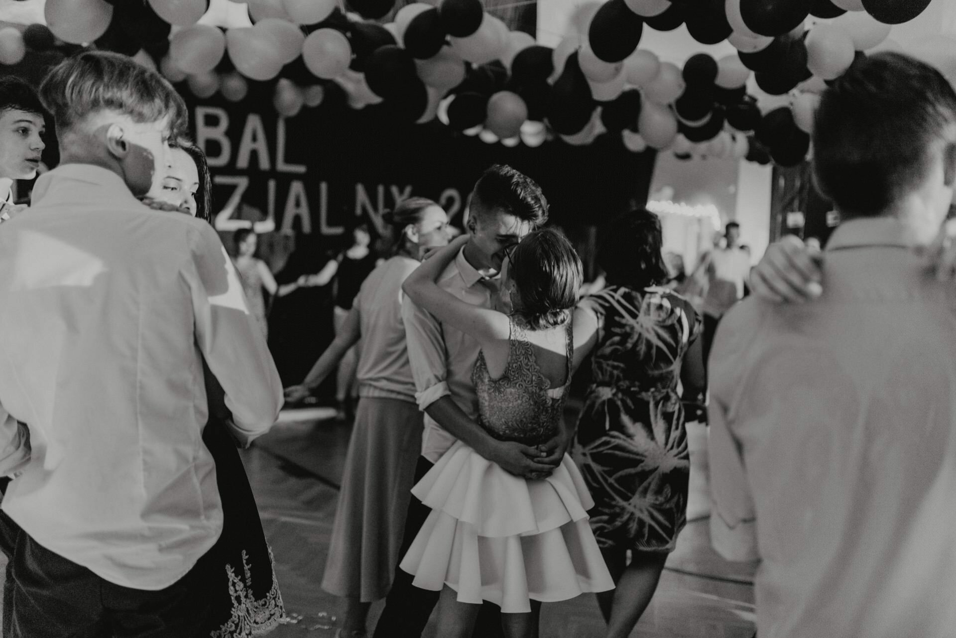 Black and white image of people dancing close together at a party or formal event. The room decorated with numerous balloons on the ceiling sets a festive mood. One couple is in the center, embracing and dancing, while others mingle in the background - a perfect example of event photography capturing timeless moments.  