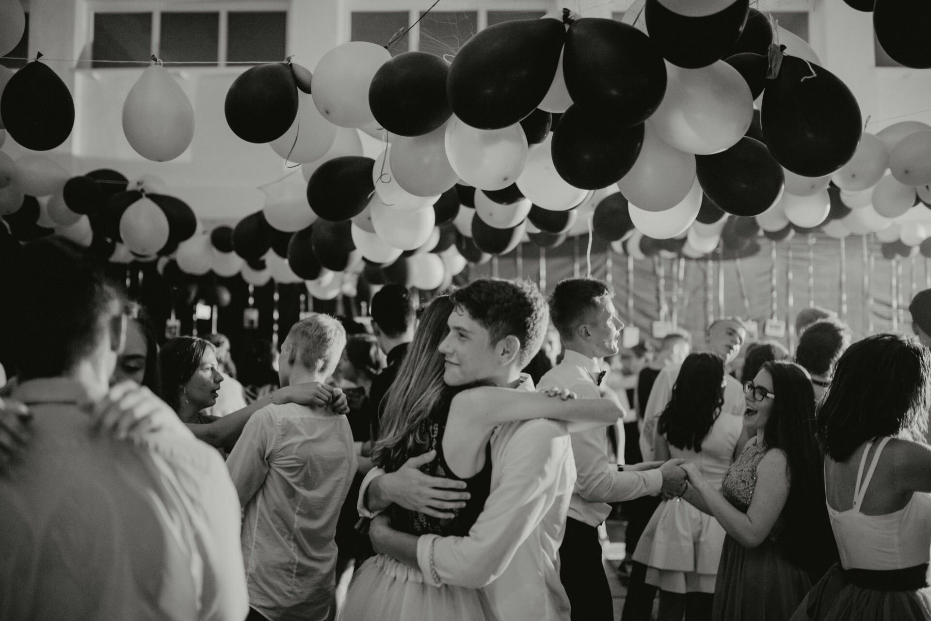 The black and white photo by event photographer Warsaw shows a crowded dance floor full of young people. Black and white balloons hang from the ceiling and couples dance close together. One couple is clearly embracing in the center of the photo, which creates a festive and lively atmosphere.  