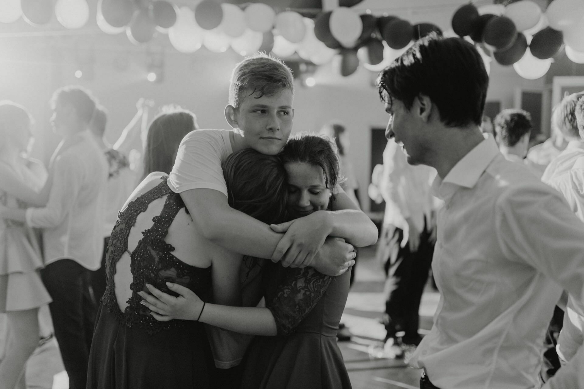 A group of young people at a dance hug each other warmly. One boy picks up two girls who embrace him tightly, while another boy stands to the right, smiling and busy in conversation. The background is decorated with streamers and balloons to capture the perfect moment for event photography.  