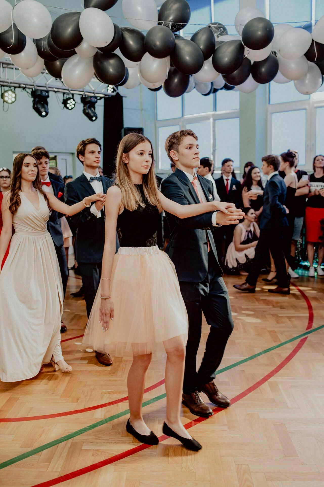 A group of young people dressed in formal attire dance in a room decorated with black and white balloons. A young woman in a beige skirt and black top dances with a young man in a suit and bow tie. The other participants stand in the background and watch, which was beautifully captured by the event photographer warszawa.  