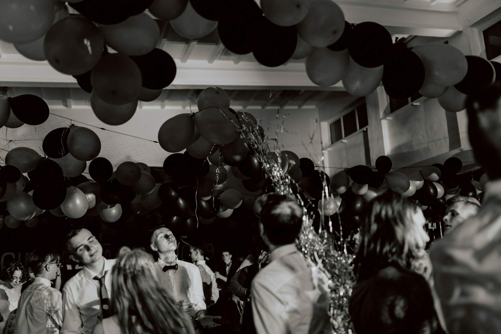 The black and white photo shows people at a lively celebration with balloons decorating the ceiling. Several people look up, some smile, while streamers float into the air, adding to the festive atmosphere. This captivating event photography perfectly captures the joyous moments.  