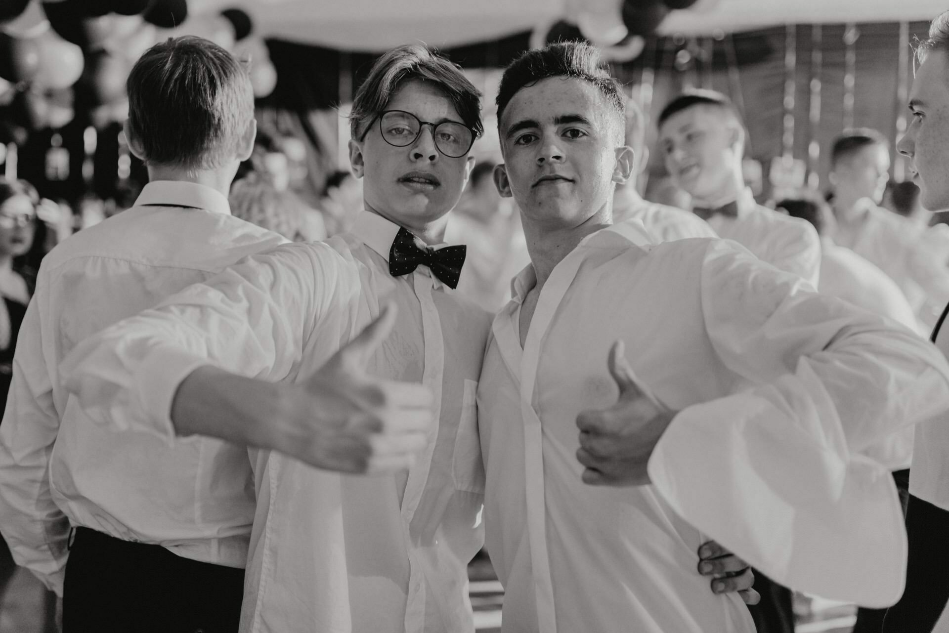 The black and white photo of two young men at a formal event captures the essence of event photography. Both are wearing white shirts; the one on the left has a bow tie and glasses. Posing, embracing each other's arms and showing thumbs up, they stand among others in similar attire.  
