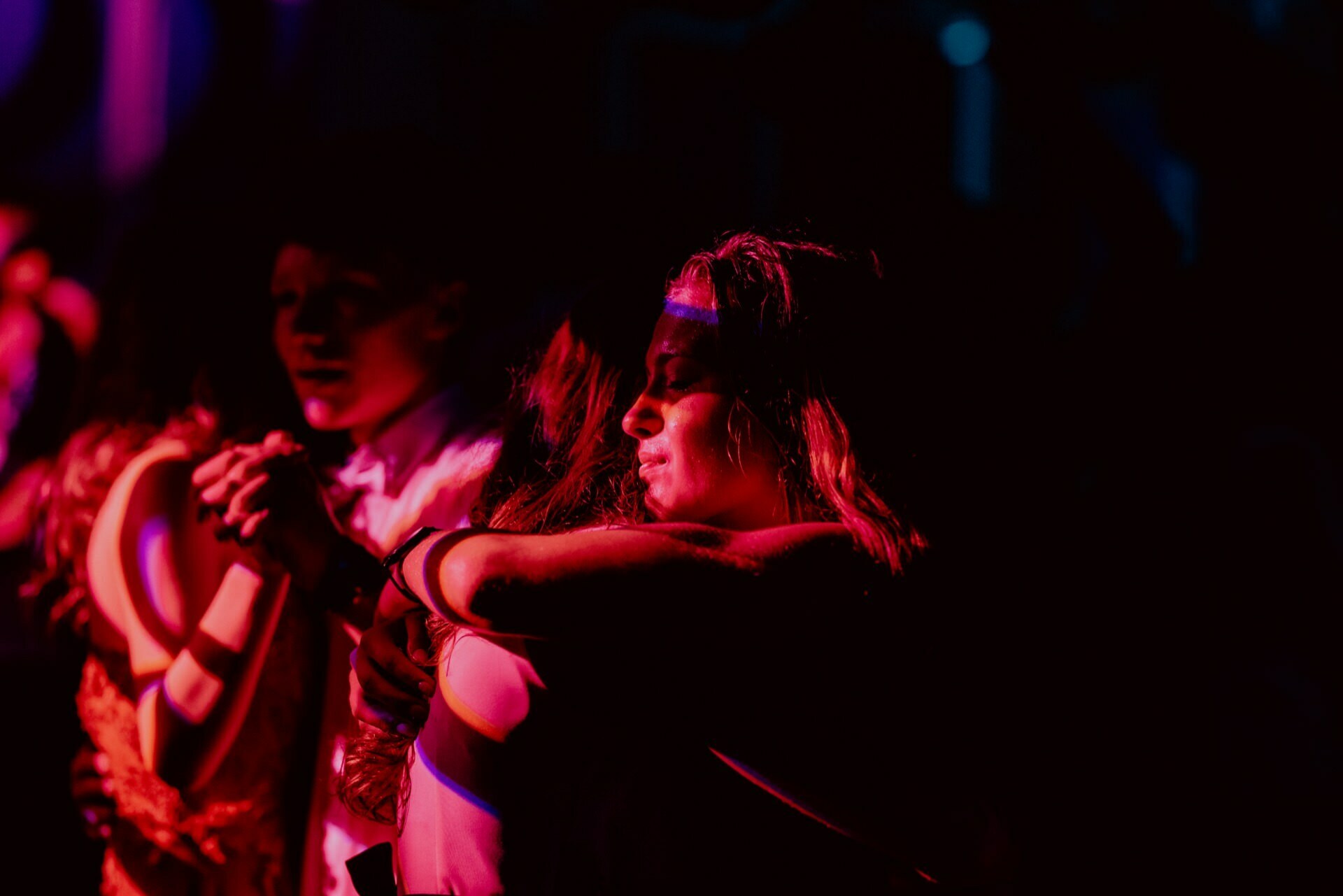 A group of people embrace in a dimly lit environment with vivid, multicolored lighting casting shadows. The expression on their faces suggests deep emotion or comfort. A scene captured by an event photographer Warsaw gives the impression of intimacy or celebration.  