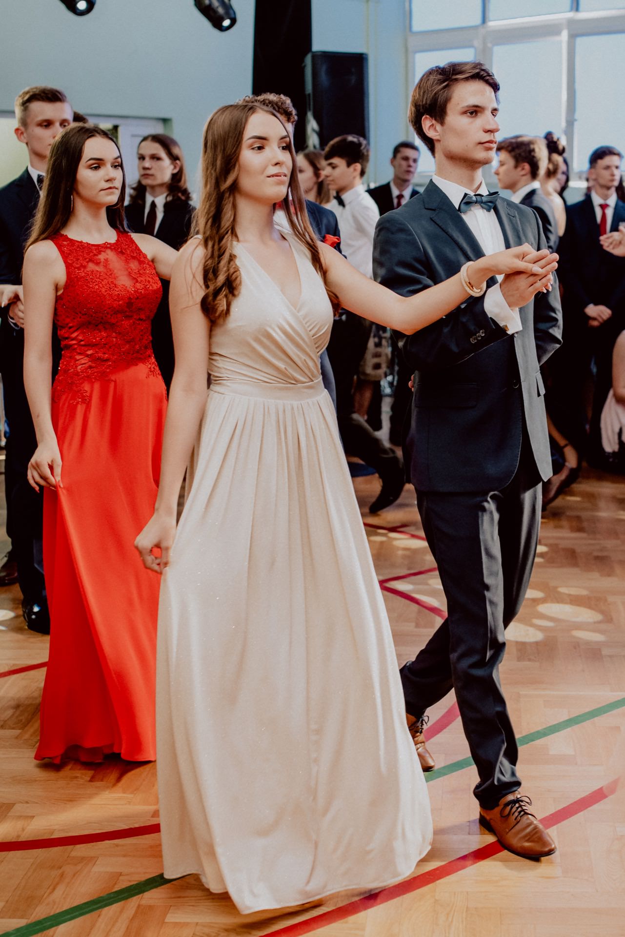 A group of young people in dance in formal attire. In the foreground, holding hands, stand a girl in a white dress and a boy in a dark suit with a bow tie. They are followed by a girl in a red dress. In the background you can see more figures, also dressed formally - perfect for event photography.   