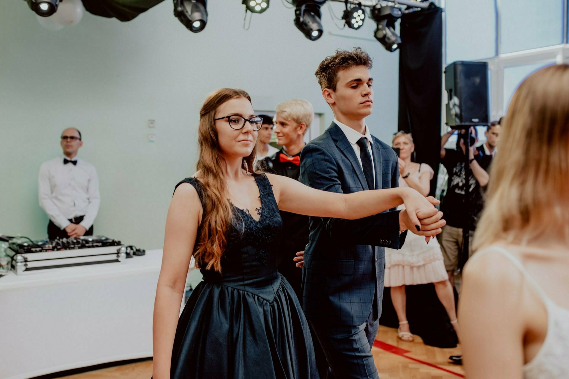 A woman in a dark dress and glasses is partnered with a man in a dark suit at an event held indoors, beautifully captured by event photography. They hold hands as they dance, while other attendees and the DJ can be seen in the background. This snapshot is part of an event photo essay.  