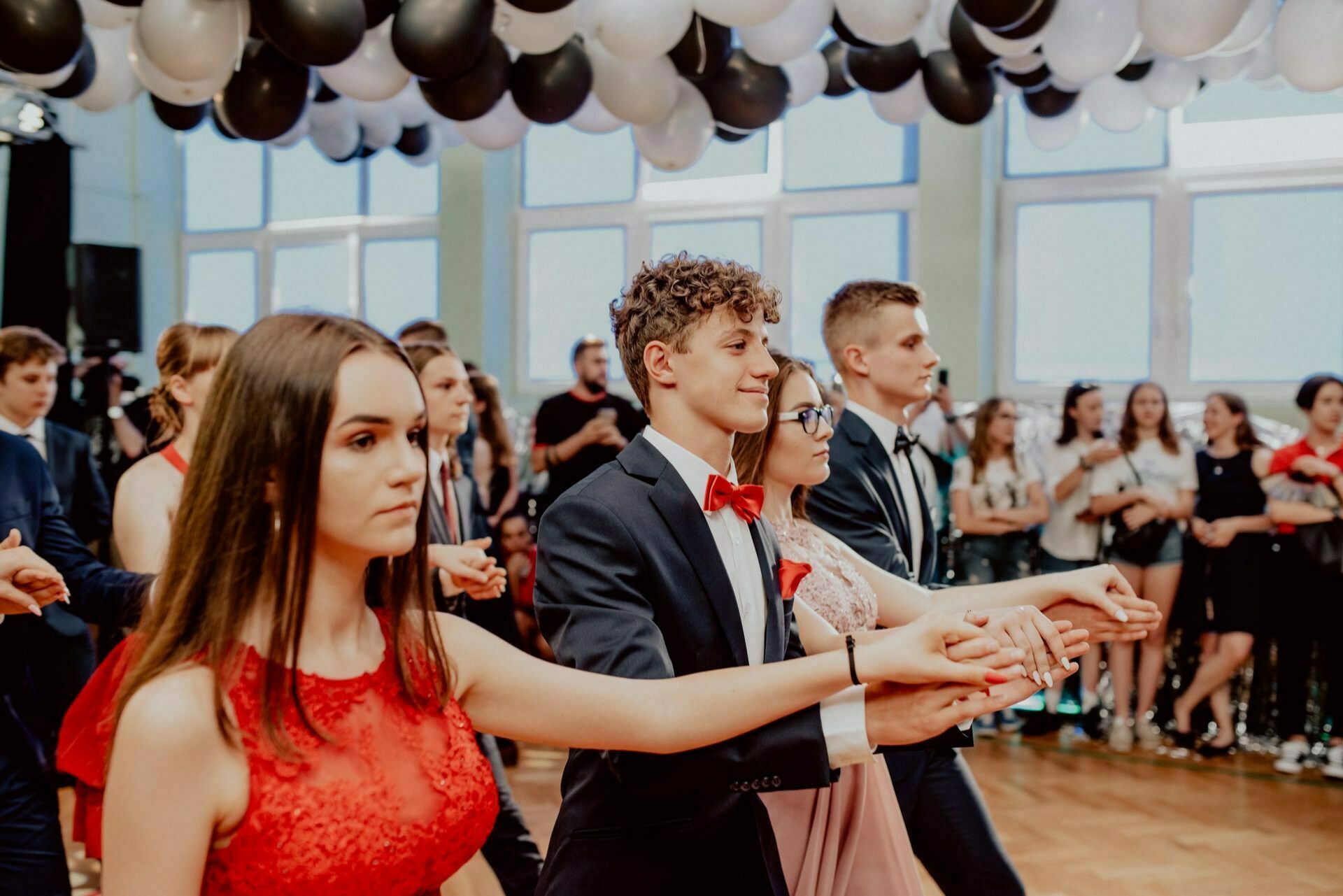 A group of young people dressed in formal attire dance in pairs in a decorated room with black and white balloons on the ceiling. They appear to be performing a choreographed routine, with the audience standing in the background, which was perfectly captured by an event photographer Warsaw who documented this vibrant event photo shoot. 