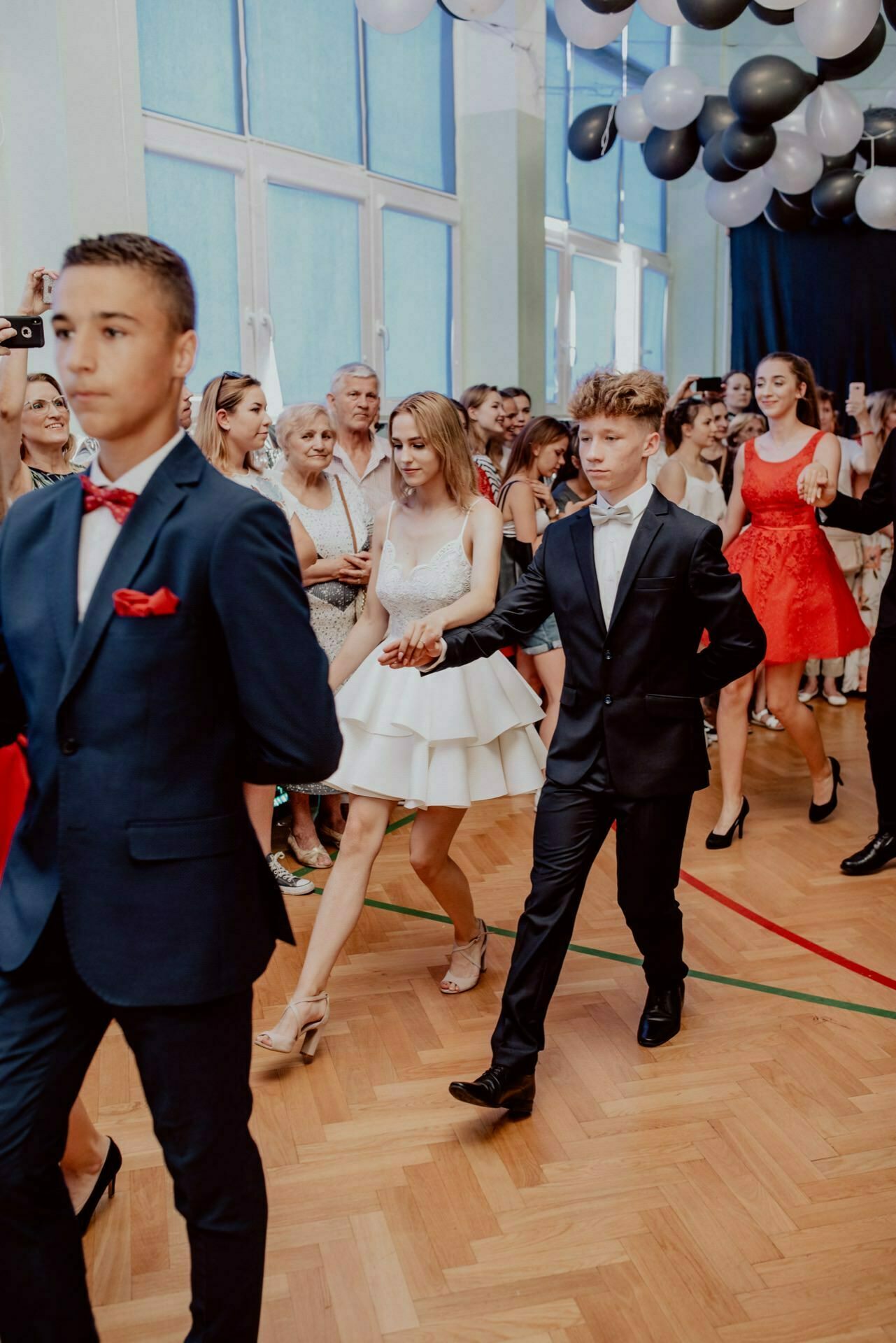 A group of young people dressed in formal attire perform a dance in a brightly lit room with a wooden floor. They are surrounded by spectators, some of whom are taking photos for event photojournalism. The dancers, holding hands and appearing focused, embody the essence of event photography.  