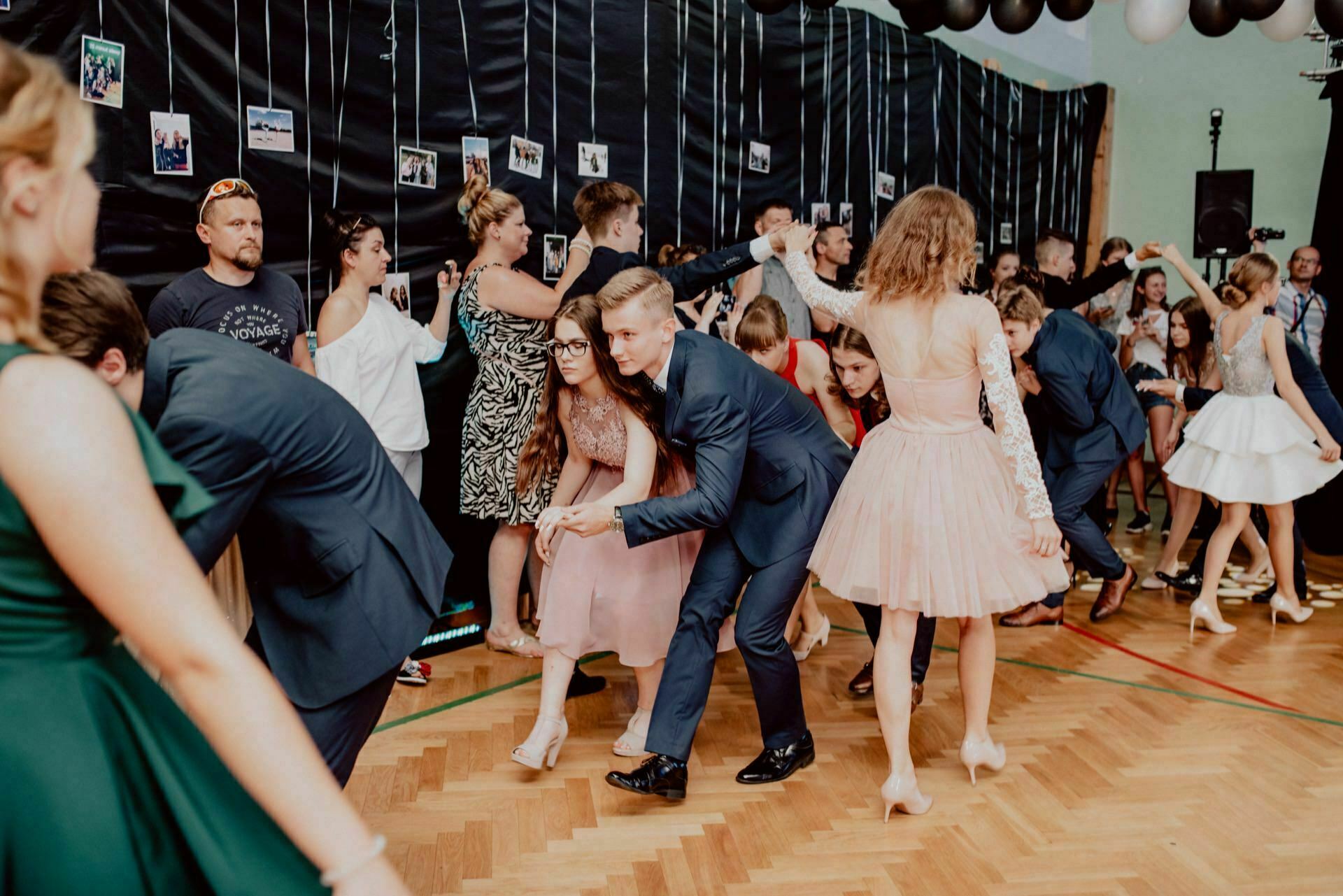 A group of people dressed in formal attire dance vigorously during a party held in a room with black wall decorations and hanging photos, creating a captivating photo-op of the events. The dancers seem to be having a good time, twirling and dipping, surrounded by spectators who are also enjoying the lively atmosphere. 