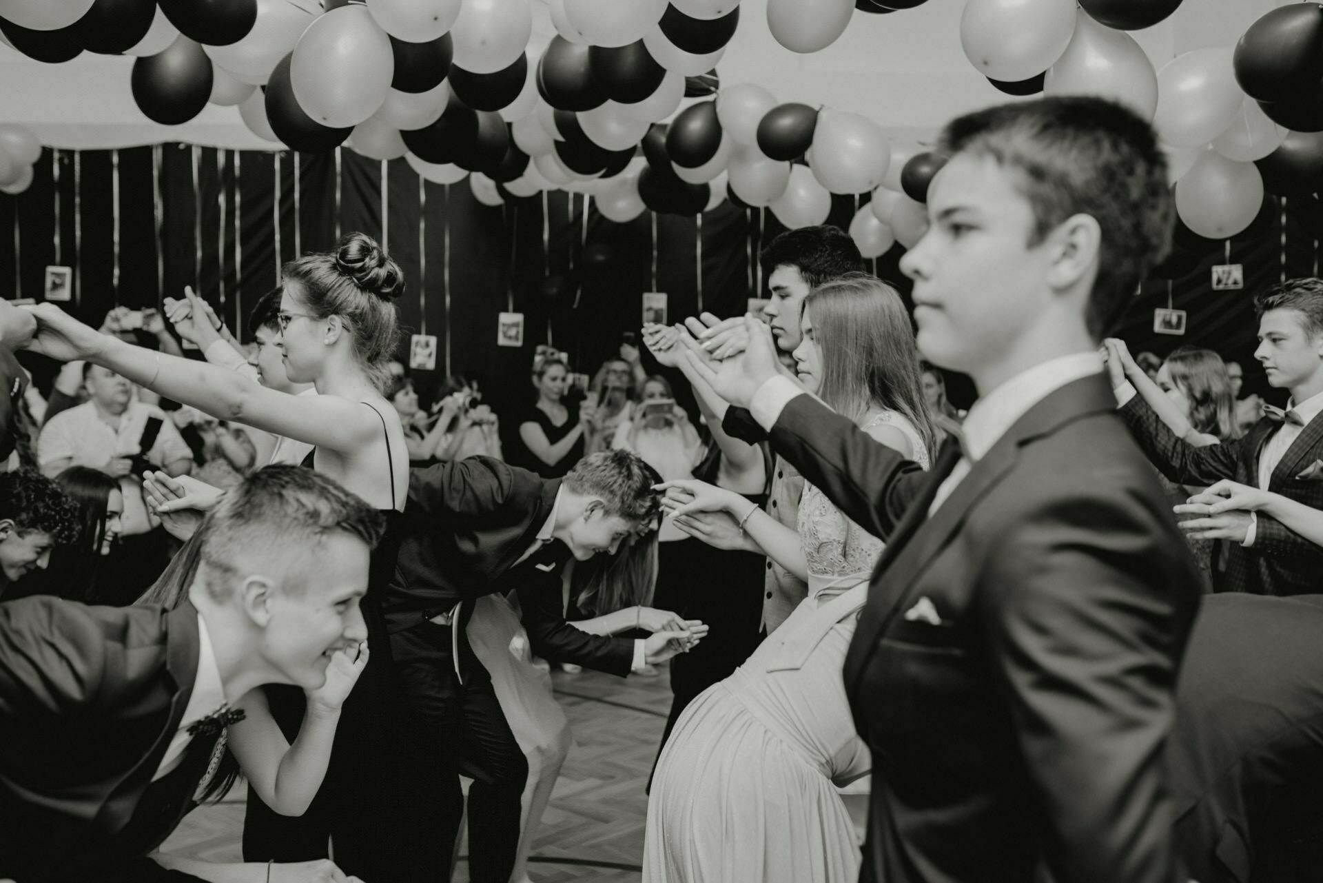 The black-and-white photo shows several young people dressed formally, participating in a dance under a canopy of balloons. The young men and women are matched in pairs and appear to be focused on their movements while the audience watches them in the background, which perfectly illustrates event photography. 