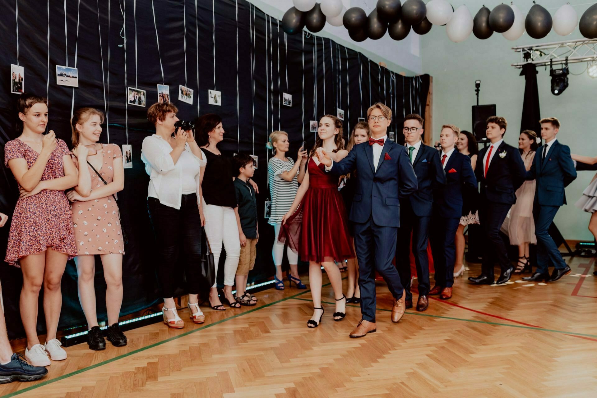 A group of people stand against a wall decorated with hanging photos, clapping and watching as a woman in a red dress and a man in a blue suit and red tie walk through the center. The room, prepared for the event by an event photographer Warsaw, has polished wooden floors and black and white balloons hanging above them. 