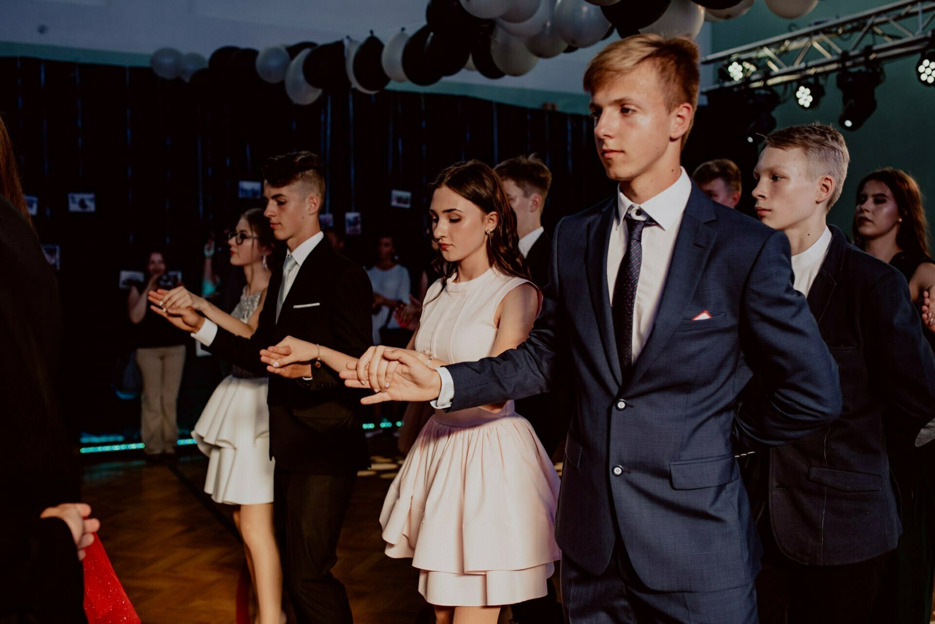 A group of young people dressed in formal attire dance in pairs at a party. The focus is on a young man in a blue suit and a young woman in a cream-colored dress. They hold hands and look serious, embodying the elegance captured in the event photography. Other couples can be seen dancing in the background.   