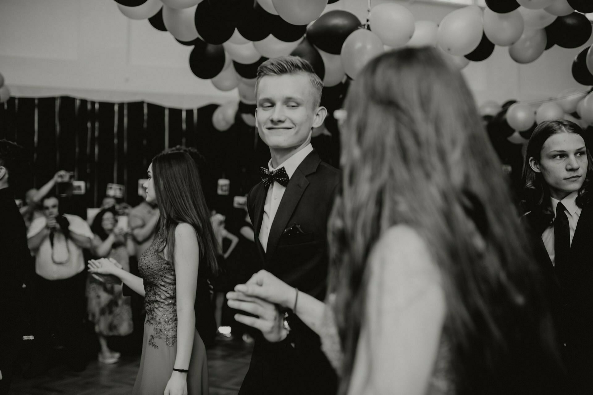 The black-and-white photograph, a stunning snapshot of the events, shows a festive dance featuring young people dressed in elegant attire. In the foreground, a young man in a suit and bow tie smiles as he dances with a young woman in a dress. Overhead, balloons and decorations are visible.  