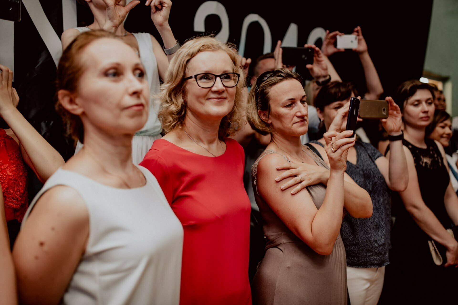 A group of people standing together, focused on an off-camera event. Some are holding phones in their hands, presumably to take photos or videos. Three women are particularly striking in the foreground, one of whom, in a red dress and glasses, smiles directly in front of her - capturing the essence of event photography.  