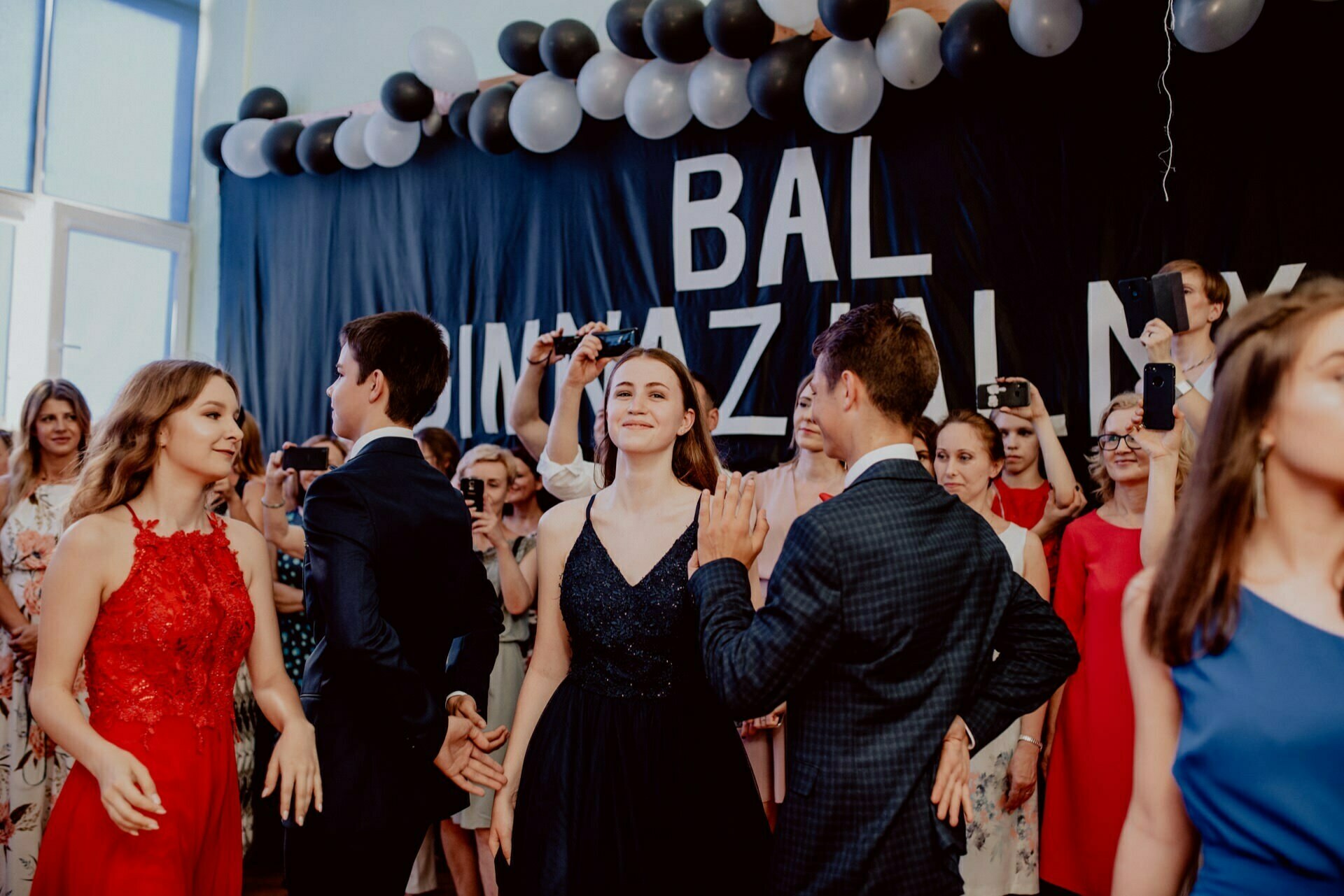 A group of young people dressed in formal attire dance at a prom or formal event. In the background is a black wall with white and silver balloons and a sign that reads "Graduation Ball." Other guests capture the moment on their phones, adding to the event photo report curated by event photographer Warsaw.  