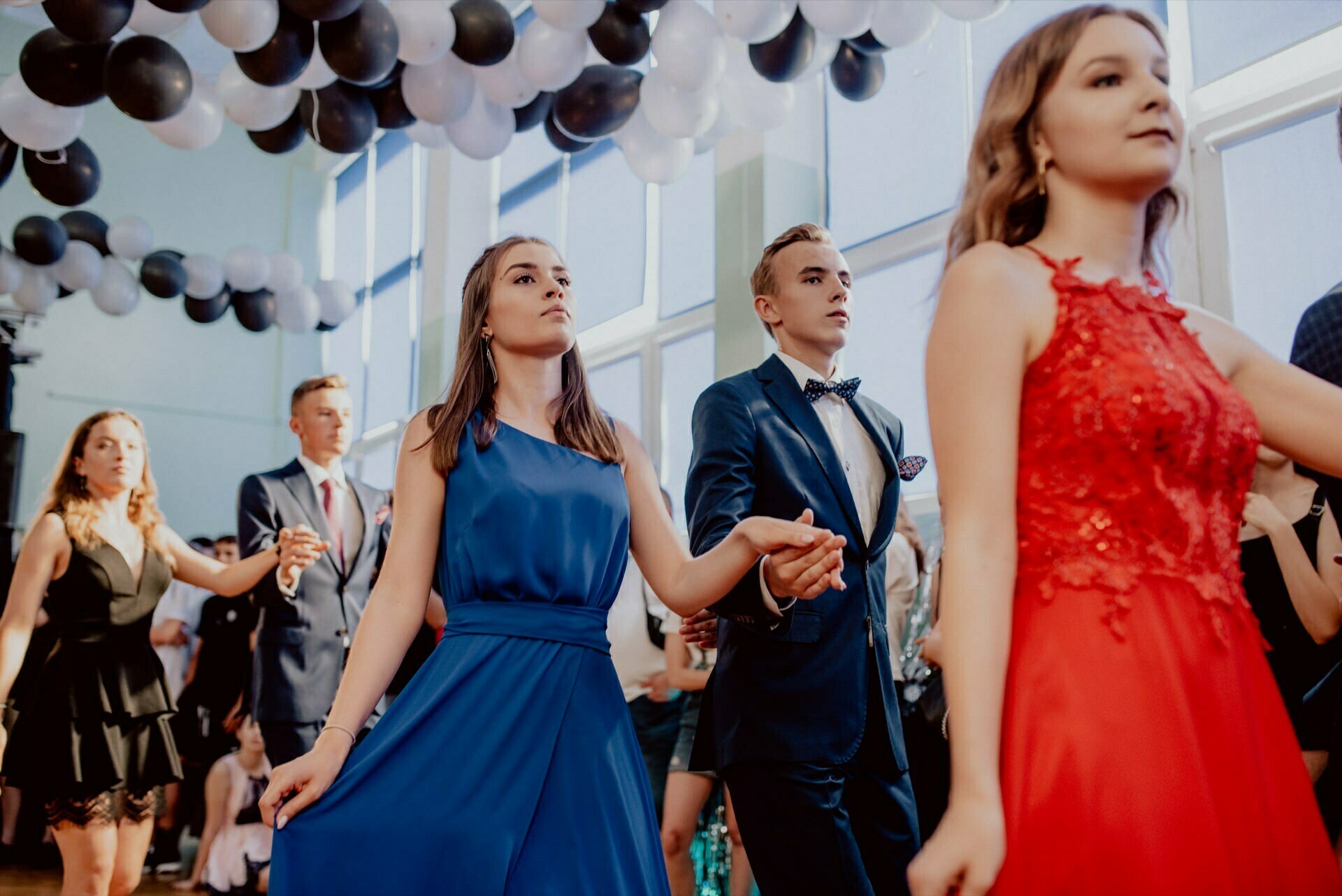 A group of young people are dancing at a party, such as a prom or formal gathering. They are dressed in elegant outfits; the young woman in the foreground is wearing a red dress, while another woman nearby is wearing a blue dress. The room, beautifully captured by event photographer Warsaw, is decorated with black and white balloons.  