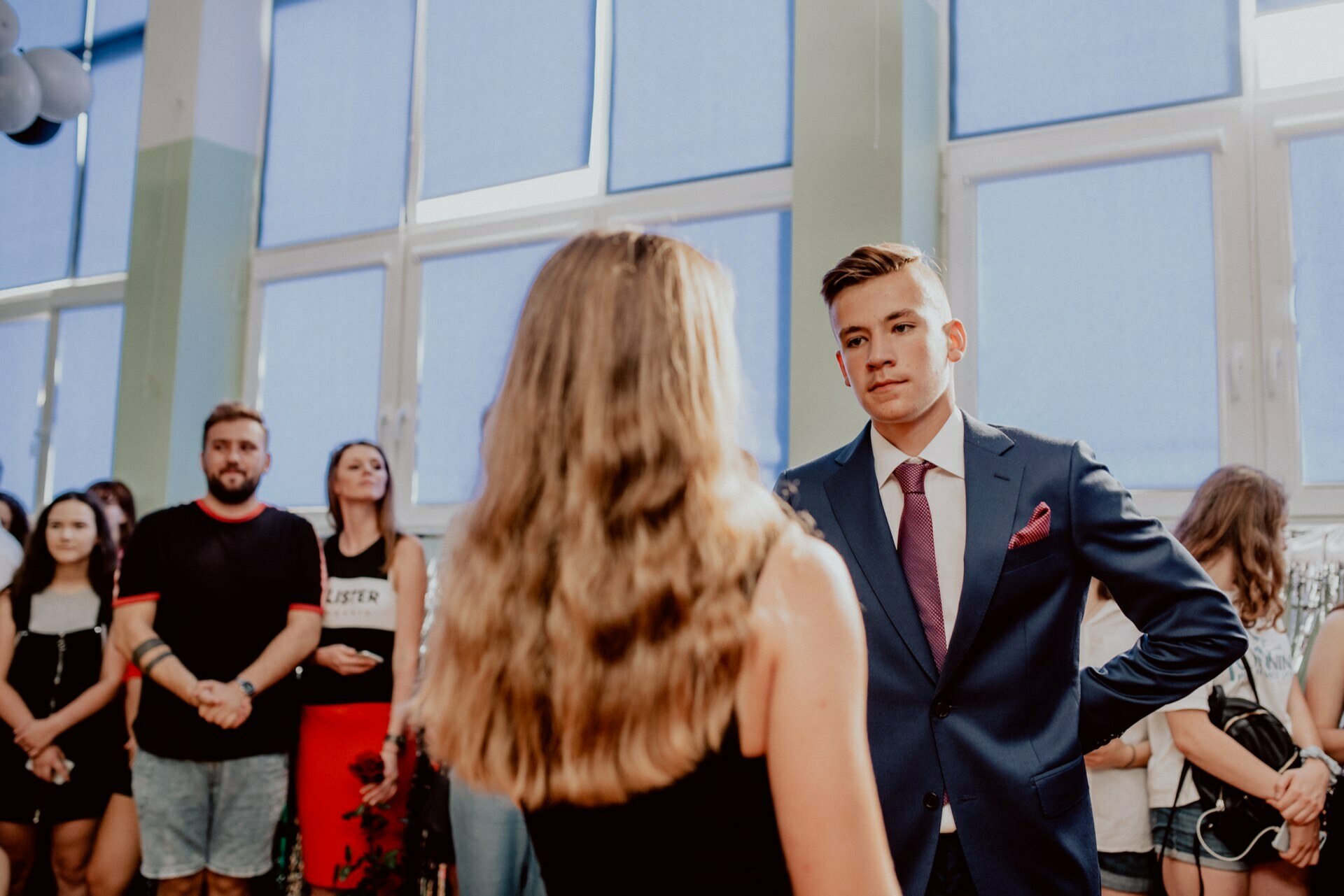 A young man in a suit and tie is facing a young woman with long hair in a black dress, in a room full of people. They look as if they are at some event or gathering, while others in casual attire watch them in the background. Behind the group are large windows with blue blinds. This scene perfectly captures the essence of event photography.   