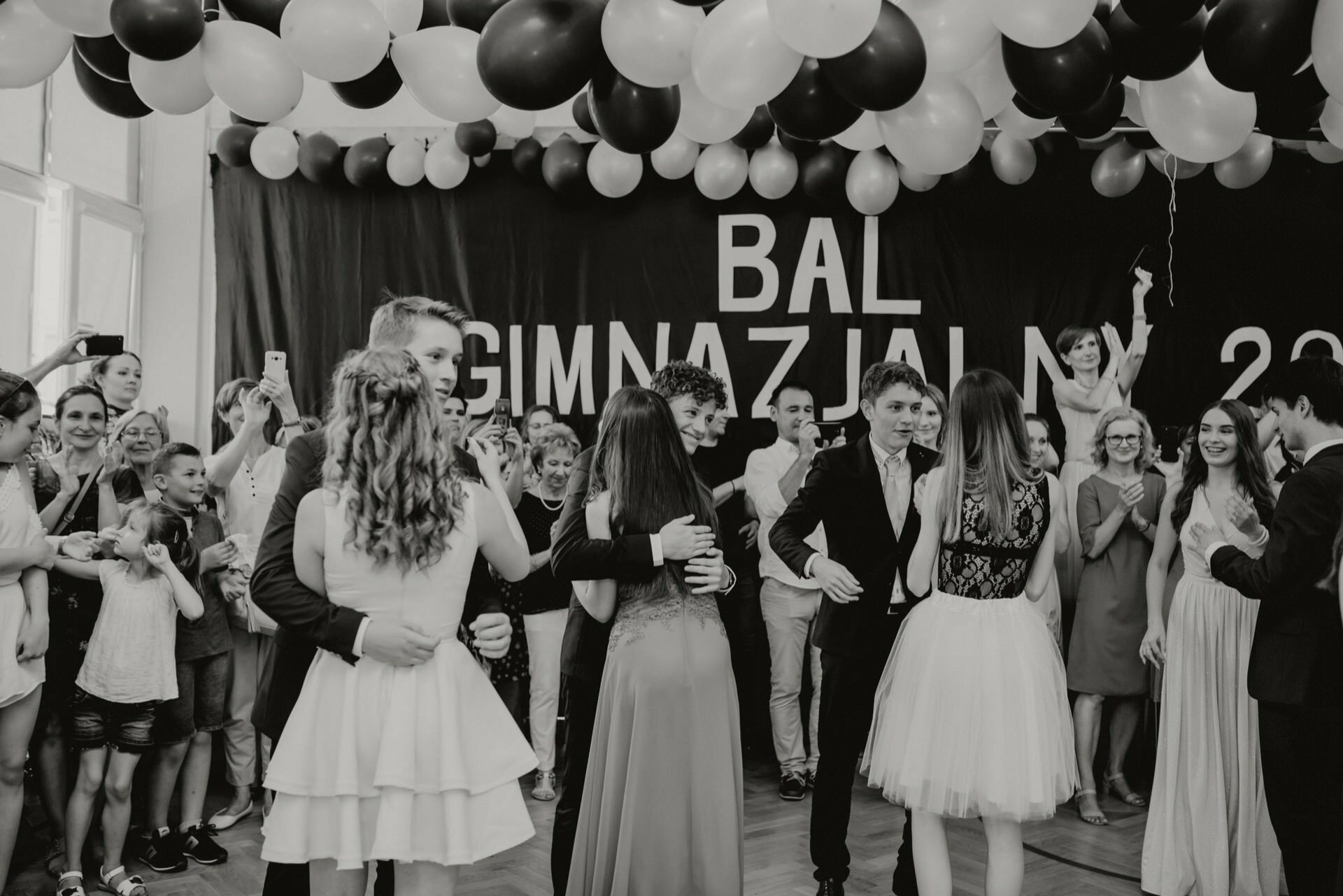 Black and white photo of a school ball. Several young couples are dancing under a ceiling decorated with many balloons. Behind them a crowd of people, including children and adults, are watching and taking pictures. A sign in the background reads "Middle School Ball 2023." This is part of an event photo report by event photographer Warsaw.    