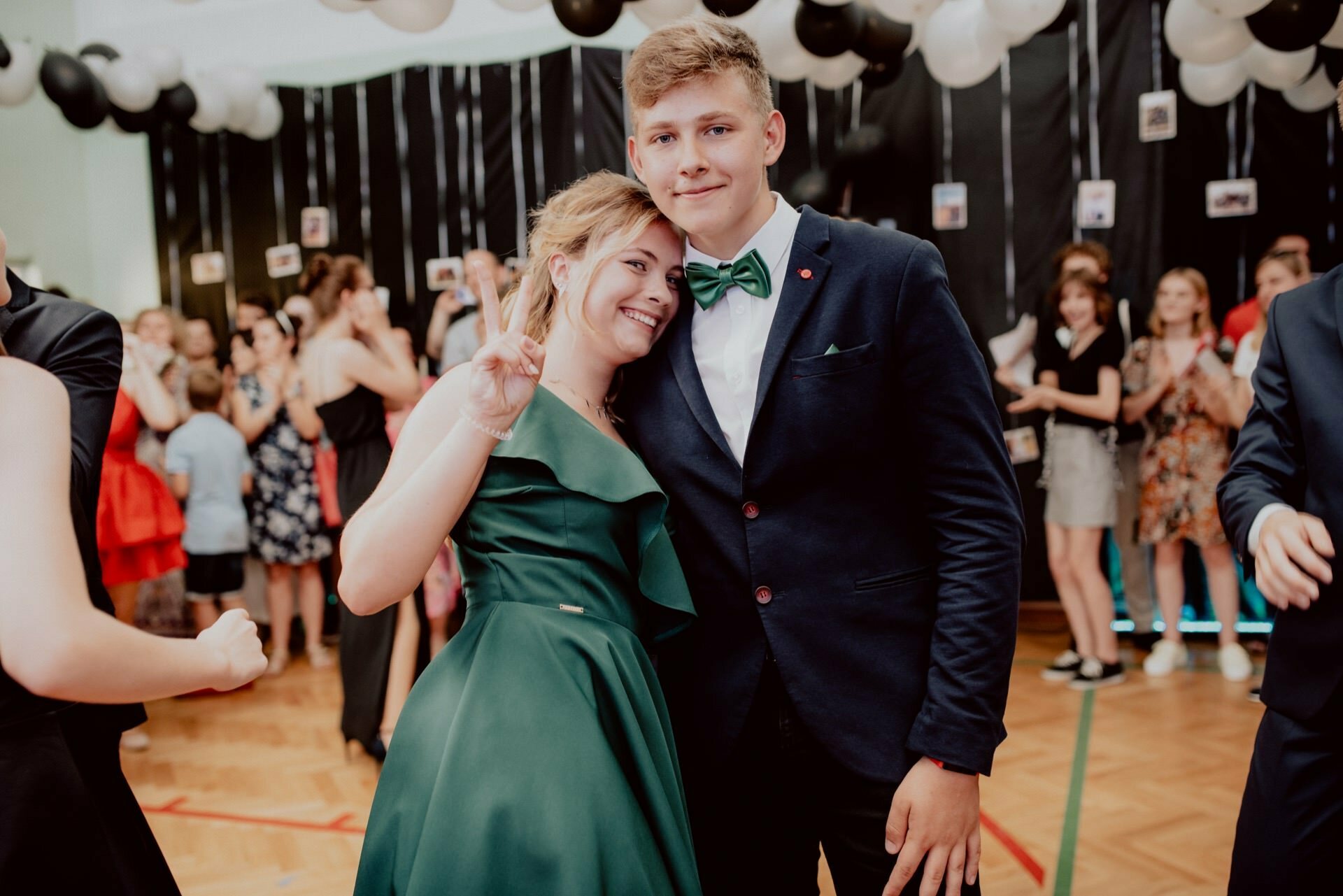 A young woman in a green dress and a young man in a dark suit with a green bow tie pose together at an official event. The woman makes a peace sign with her hand. Other people mingle in the background, and black and white balloons hang from the ceiling, capturing the perfect event photography moment.  