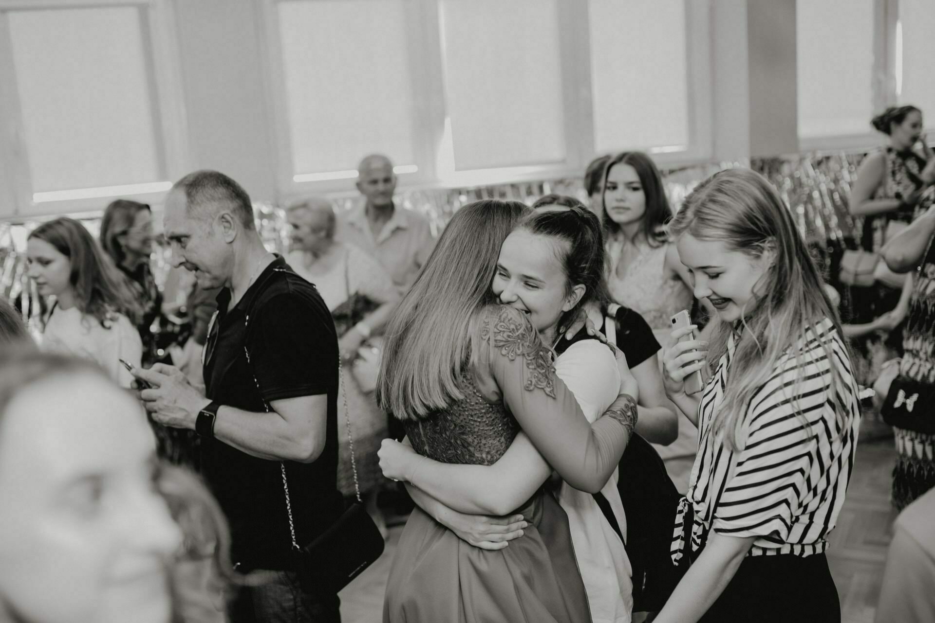 The black and white photo of the crowded room captures the essence of event photography. In the middle, two women are hugging, smiling. The people around them are engrossed in conversation, and some have cameras, documenting the photojournalism of the events. The room has large windows in the background.   