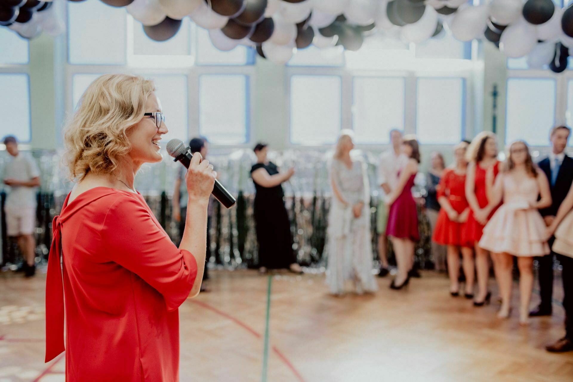 A woman in a red dress and glasses speaks into a microphone at a room party. A group of formally dressed people stand in the background. The ceiling is decorated with black and white balloons, and large windows let in natural light, capturing the perfect scene for event photography.  