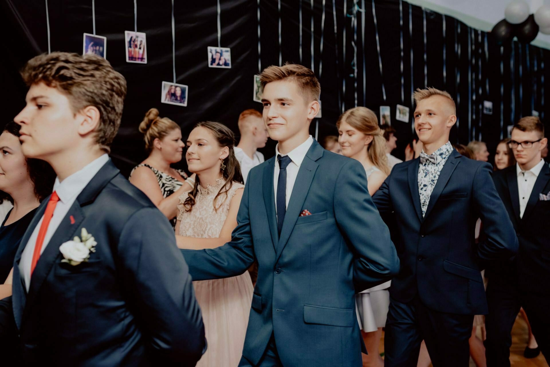 A group of young people dressed in formal attire dance in pairs at a party, against a backdrop of black walls and hanging photos, elegantly captured by an event photographer Warsaw. The men are dressed in suits and ties, while the women wear elegant dresses. The atmosphere seems joyful and festive.  